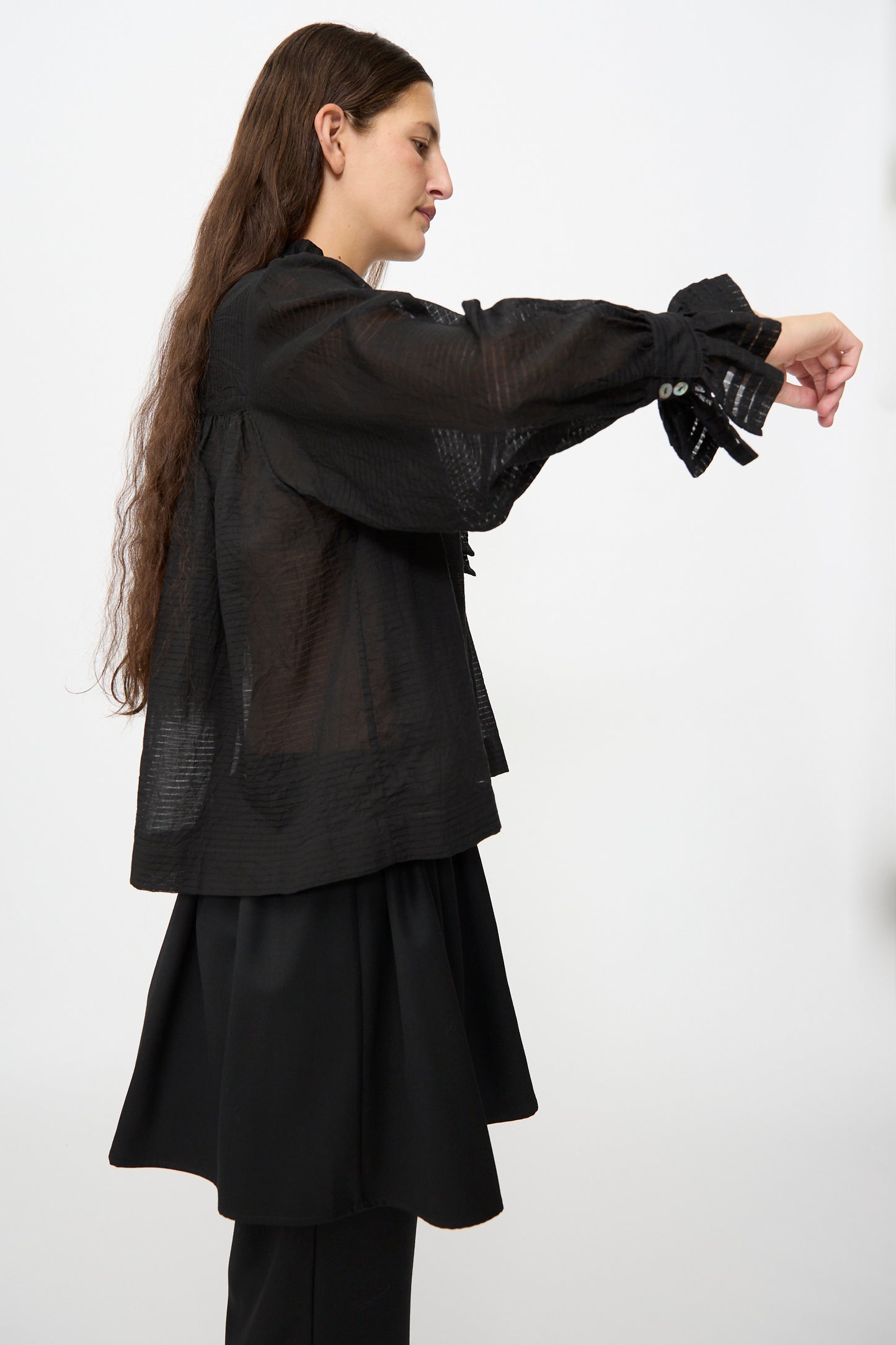 A person in profile wears the Cawley Japanese Sheer Stripe Cotton Sophie Shirt in Midnight, a loose blouse with wide sleeves, layered over a black skirt and pants, standing against a plain white background.