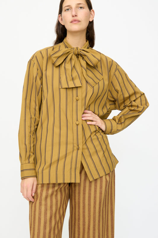 A person wears the Cawley Japanese Silk Cotton Hattie Shirt in mustard and brown stripe, featuring long sleeves and a large neck bow, paired with striped pants, against a plain background. Made in London.