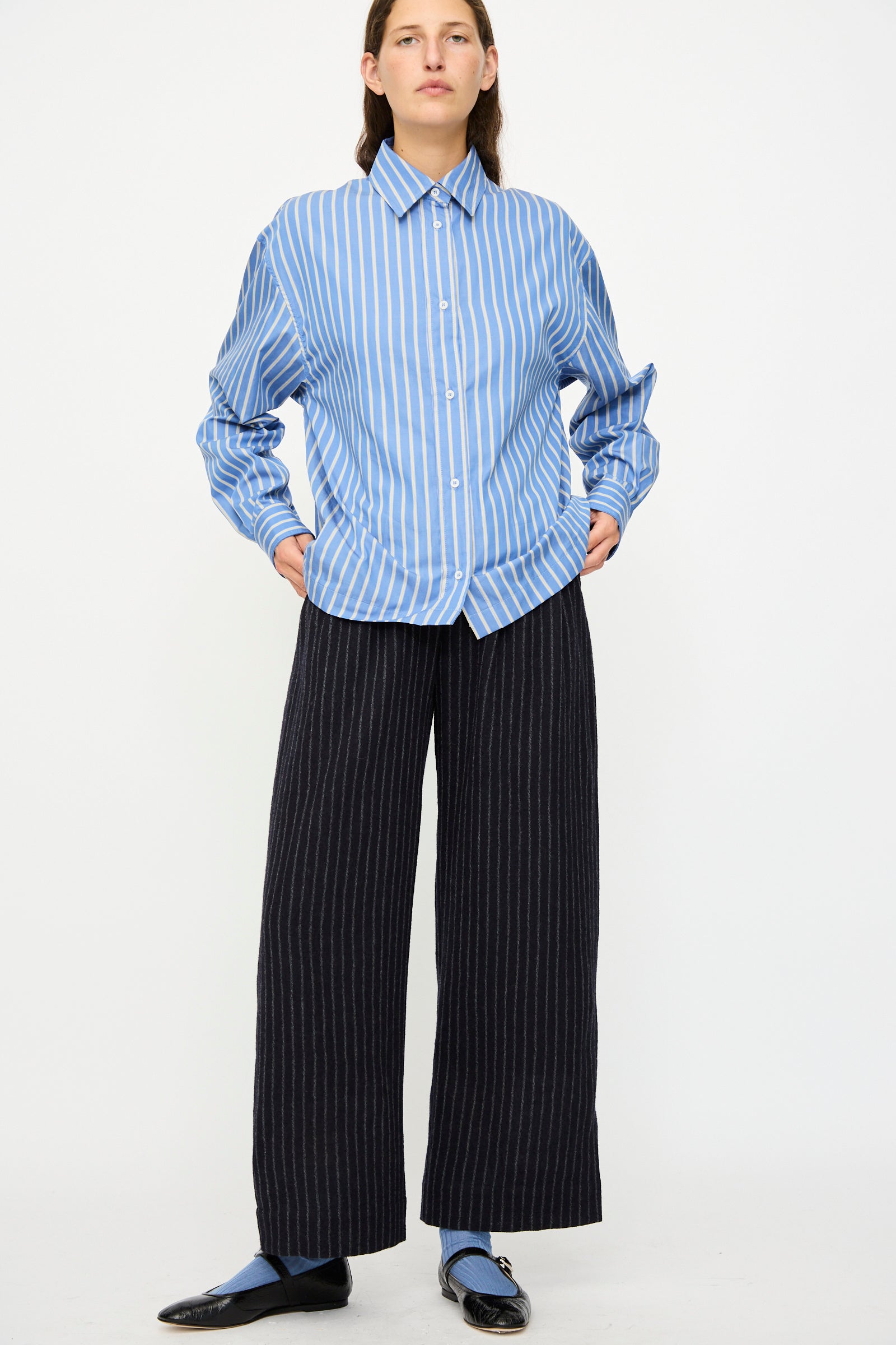 Person standing against a white background wearing a blue and white striped button-up shirt, Cawley Japanese Striped Boucle Mara Trouser in Navy and Grey, and black shoes with blue socks.