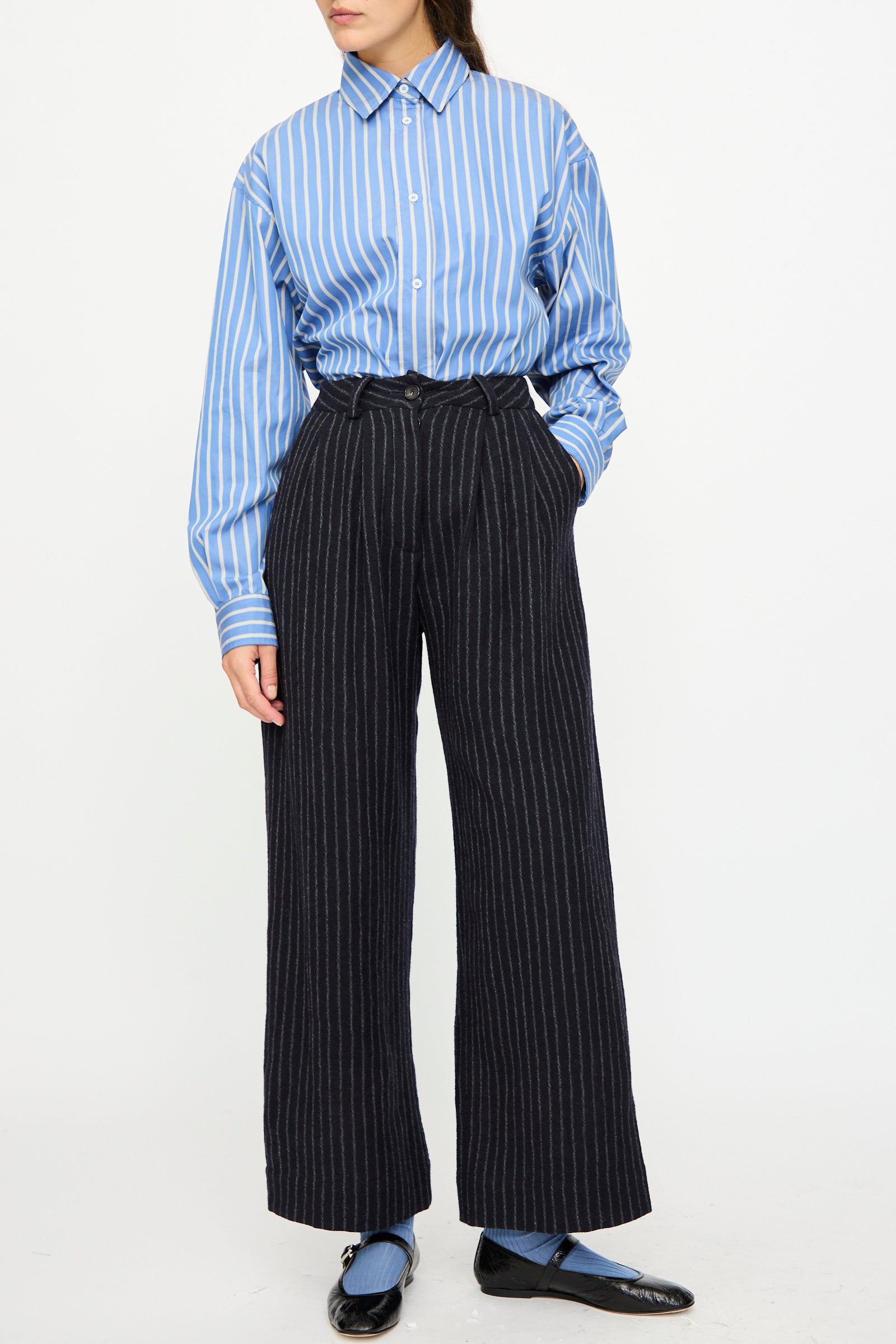 A person wearing a blue and white striped shirt tucked into Cawley’s Japanese Striped Boucle Mara Trouser in Navy and Grey, paired with black shoes and blue socks.
