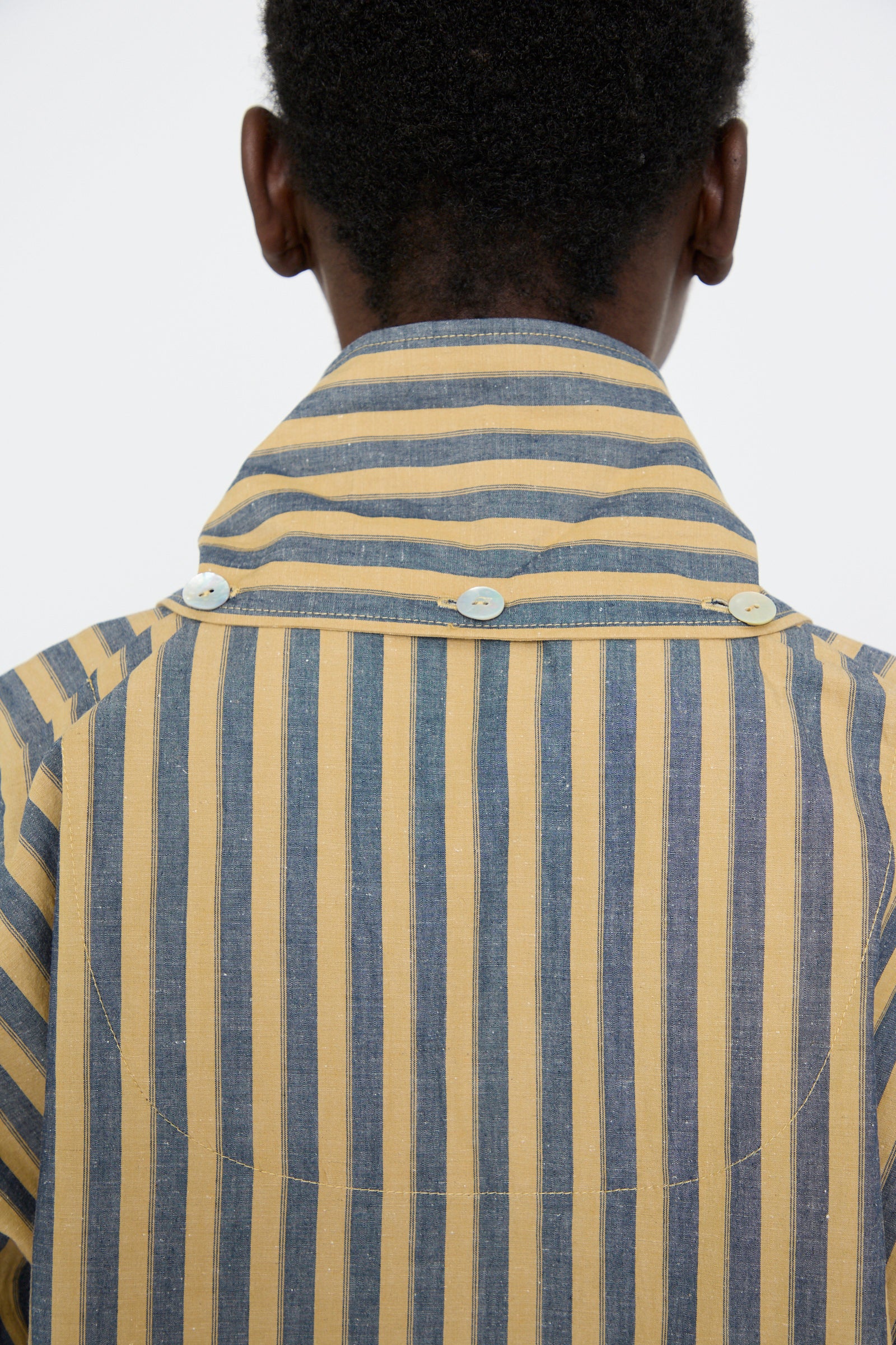 A person is wearing a relaxed fit Dorothy Shirt by Cawley, featuring mustard and navy Japanese striped cotton with long sleeves and back buttons, facing away.