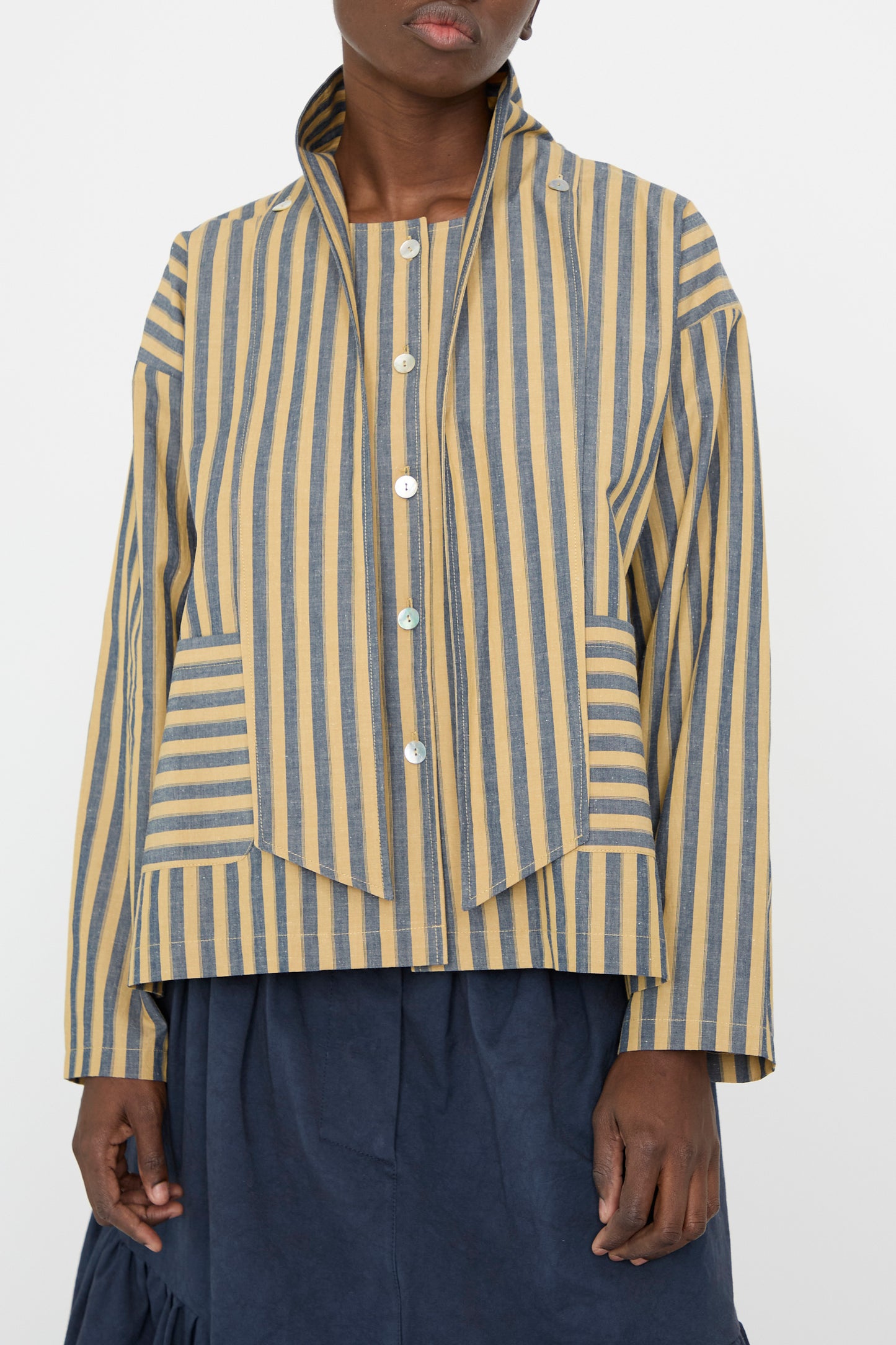 A person drapes the Cawley Japanese Striped Cotton Dorothy Shirt in Mustard and Navy, with white buttons and a high collar, over a dark blue skirt for a relaxed fit.