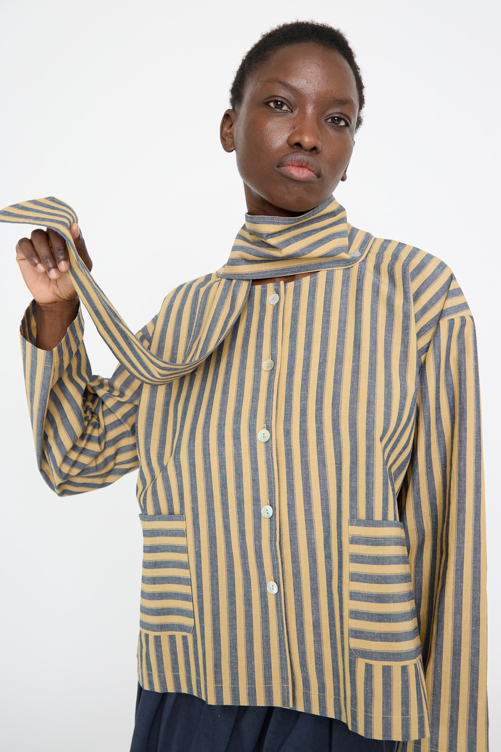 A person wearing Cawley's Japanese Striped Cotton Dorothy Shirt in Mustard and Navy holds one end of the long collar against a white backdrop. 