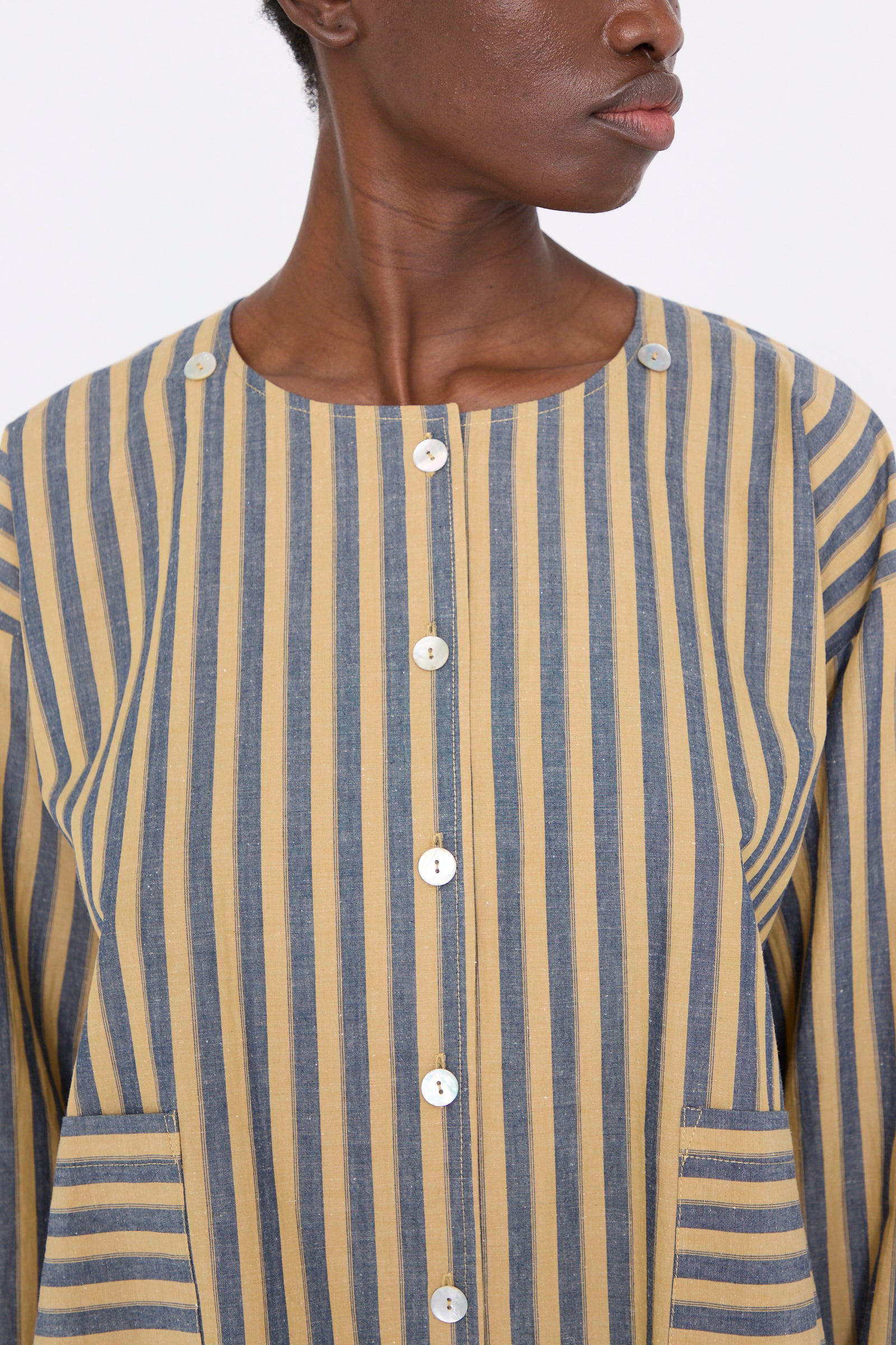 A close-up of a model is wearing the Cawley Japanese Striped Cotton Dorothy Shirt in Mustard and Navy, a relaxed-fit long sleeve shirt featuring yellow and blue stripes with two pockets.