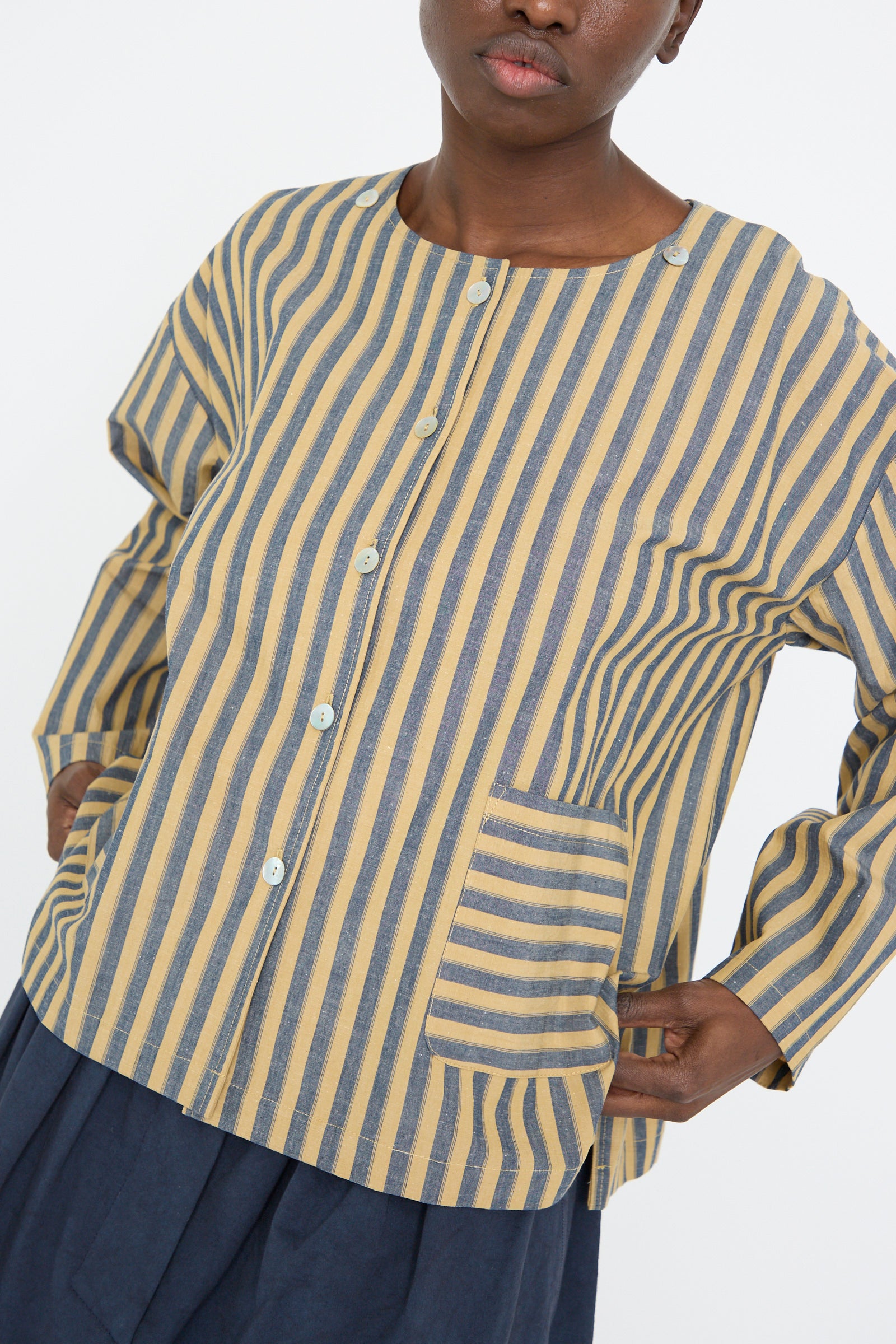 A person wearing the Cawley Japanese Striped Cotton Dorothy Shirt in Mustard and Navy, featuring a relaxed fit, long sleeves, buttons, and a large front pocket, paired with a dark blue skirt.