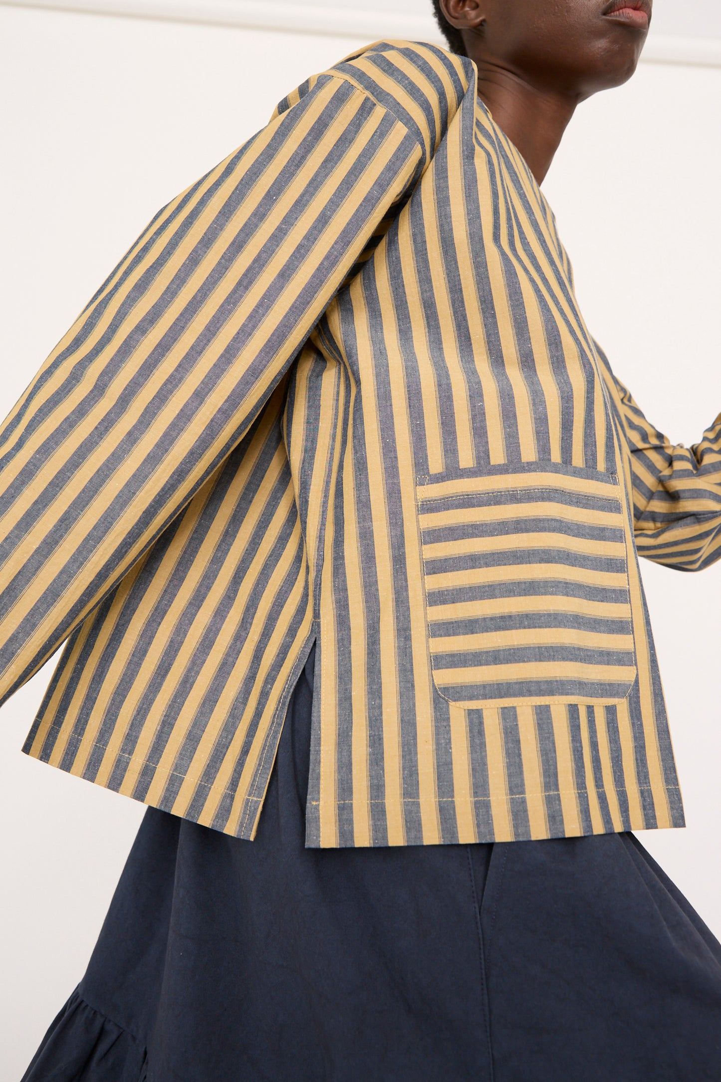 A model walks to the right, wearing Cawley's relaxed fit ensemble features the Japanese Striped Cotton Dorothy Shirt in Mustard and Navy with wide sleeves and a pocket, paired with a dark skirt.