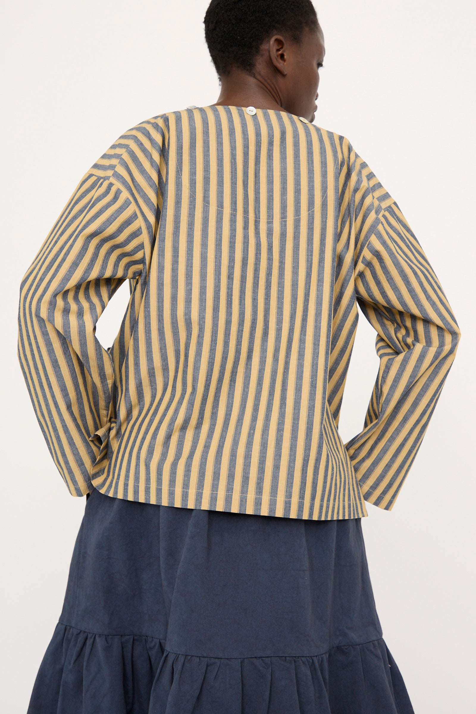 A person is photographed from the back wearing Cawley's Japanese Striped Cotton Dorothy Shirt in Mustard and Navy, featuring a relaxed fit and long sleeves, paired with a dark blue tiered skirt.