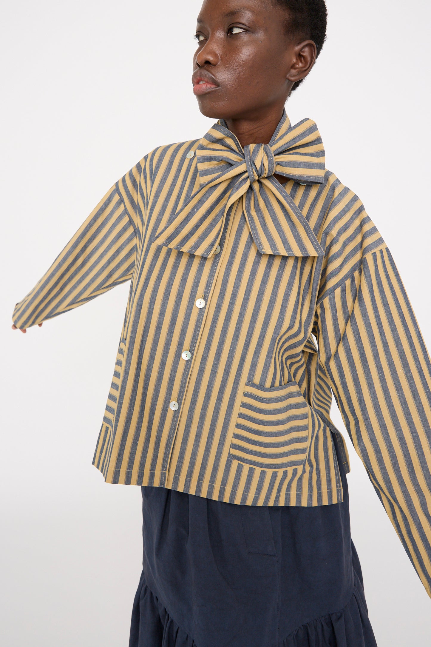 A person in a Japanese striped cotton Dorothy shirt in mustard and navy by Cawley, featuring a large bow, with arms outstretched, wearing a dark skirt against a plain backdrop.