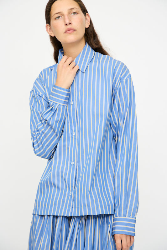 A person wears the Cawley Japanese Striped Dennis Shirt in Blue Dandelion Stripe, featuring blue and white vertical stripes, paired with a matching skirt, standing against a plain light background.