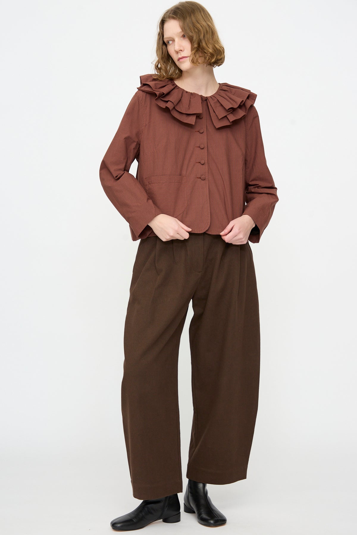 A person stands against a plain white background, wearing a brown ruffled-collar blouse, wide brown pants, and black boots, layered with the Cawley Japanese Typewriter Cotton Rowan Jacket in Burgundy Stripe for added texture.
