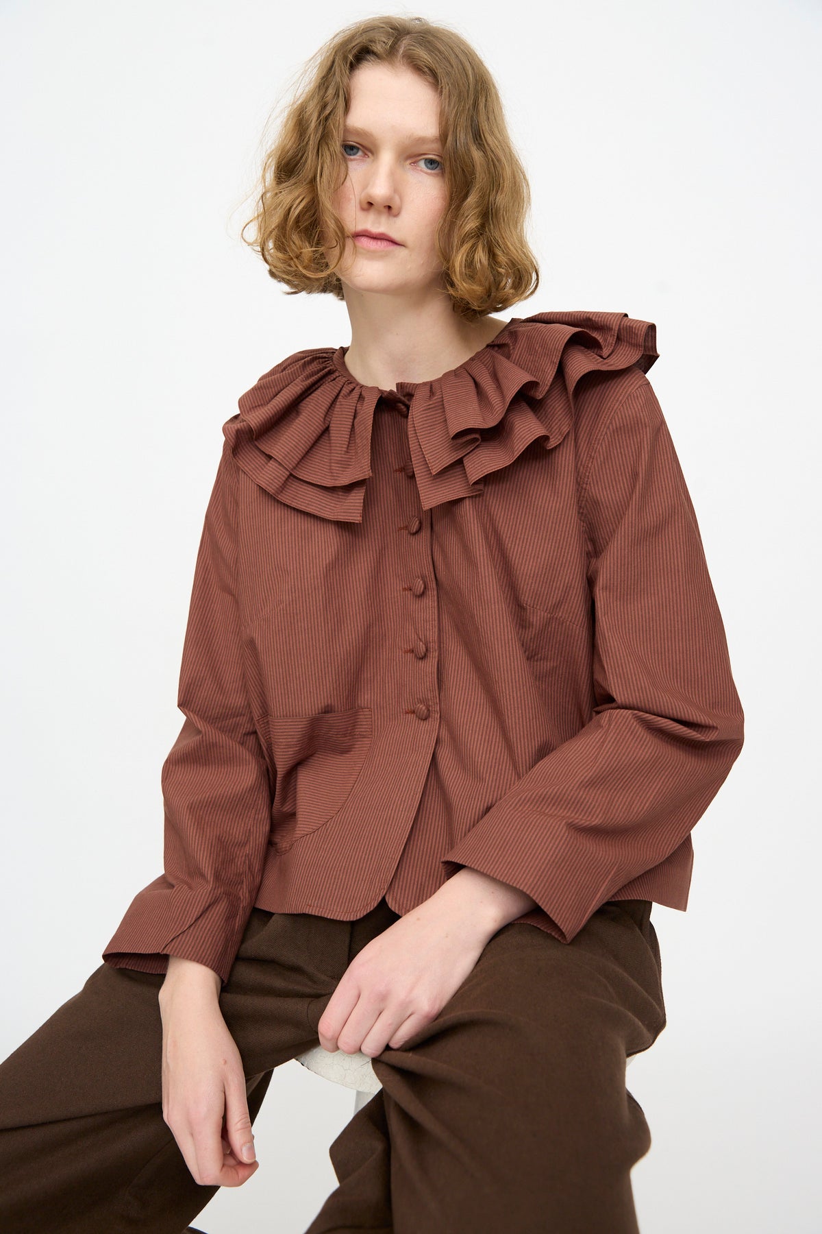 A person with wavy, shoulder-length hair wears the Cawley Japanese Typewriter Cotton Rowan Jacket in Burgundy Stripe layered over a brown ruffled blouse with buttons and brown pants, seated against a plain white background.
