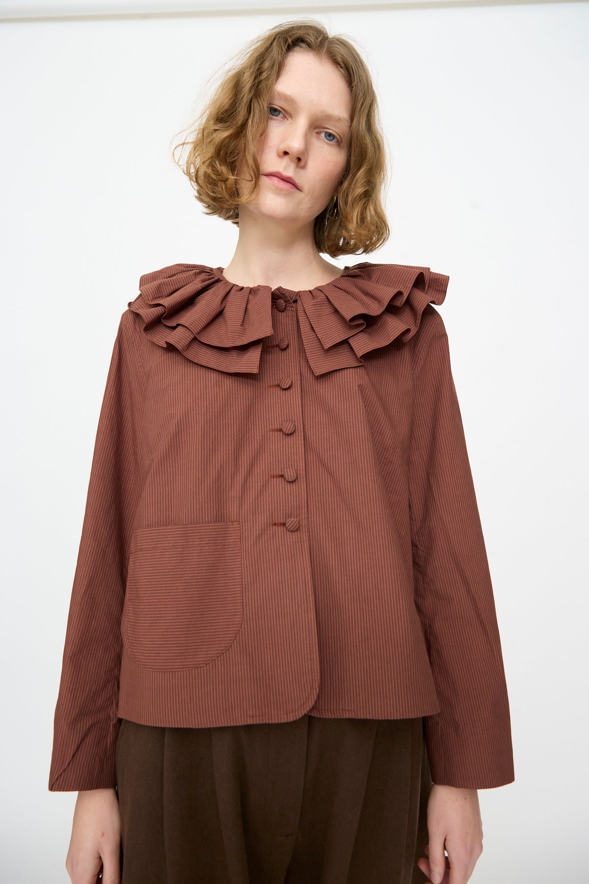 A person is wearing the Cawley Japanese Typewriter Cotton Rowan Jacket in Burgundy Stripe, featuring long sleeves and an elegant ruffled collar that adds a touch of vintage charm, paired with matching pants, and standing against a plain white background.
