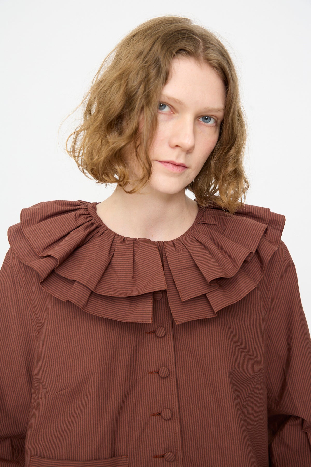 A person with wavy, chin-length hair wears the Cawley Japanese Typewriter Cotton Rowan Jacket in Burgundy Stripe, a brown pinstriped piece featuring a large, layered ruffled collar.