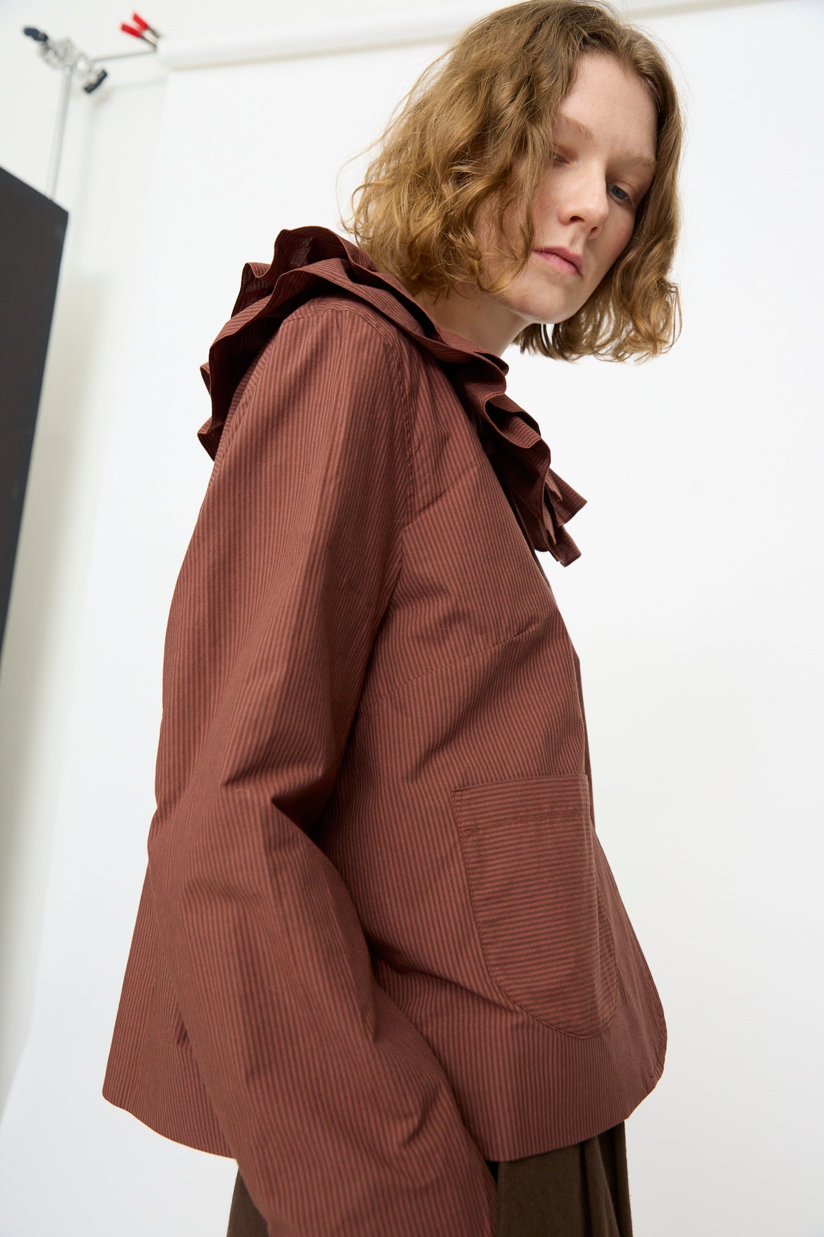 A person with wavy, shoulder-length hair is wearing the Cawley Japanese Typewriter Cotton Rowan Jacket in Burgundy Stripe over a brown, long-sleeved top with a ruffled collar and front pocket, standing against a plain background.