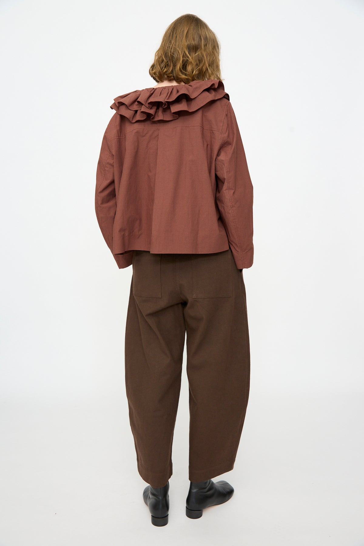 A person stands with their back to the camera, wearing the Cawley Japanese Typewriter Cotton Rowan Jacket in Burgundy Stripe, paired with loose brown pants and black shoes, against a plain white background.