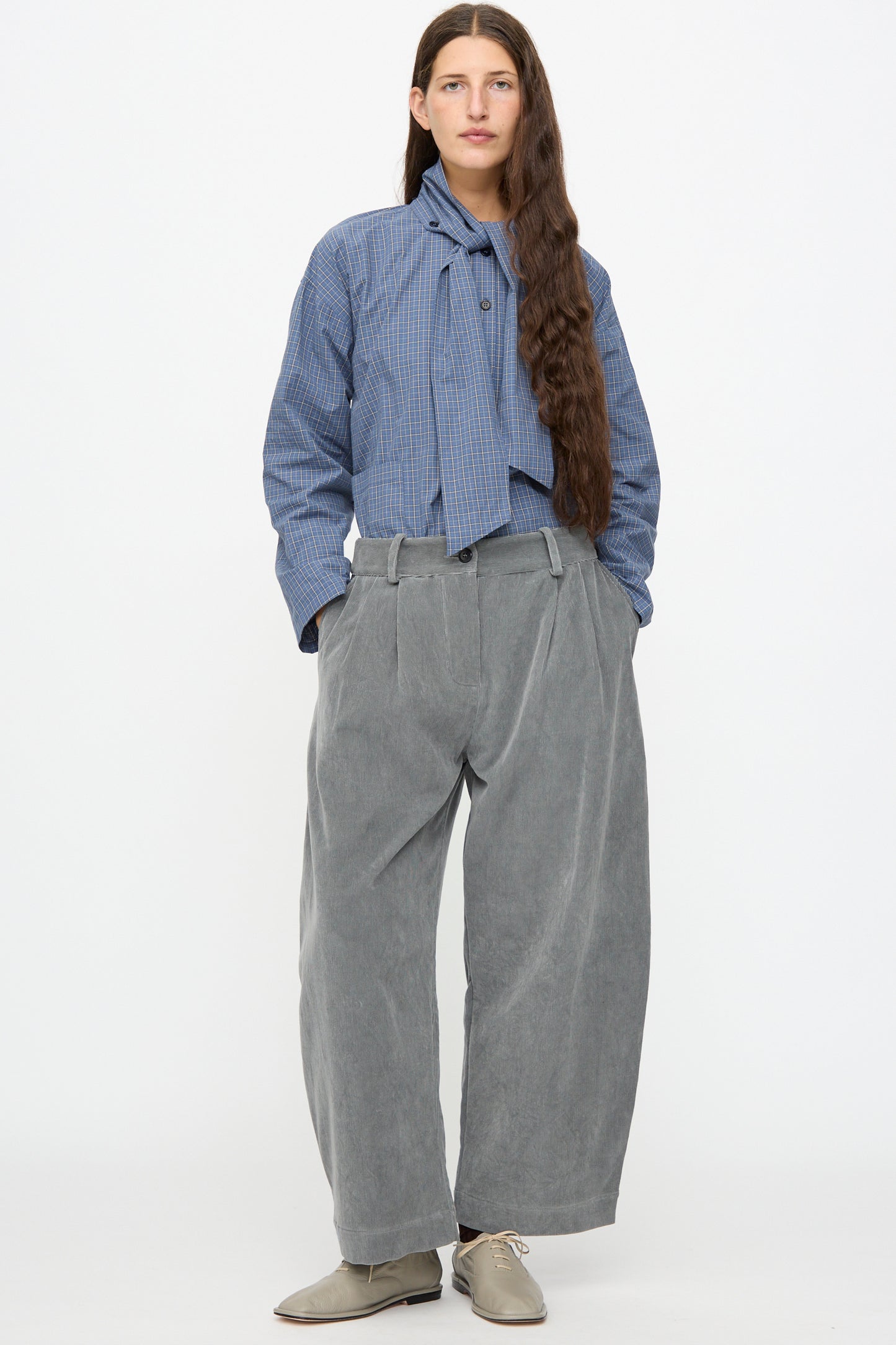 A person stands against a white background wearing a blue checked blouse with a necktie detail, paired with Cawley’s Japanese Wale Cotton Cord Robyn Trouser in Fossil and light gray shoes.