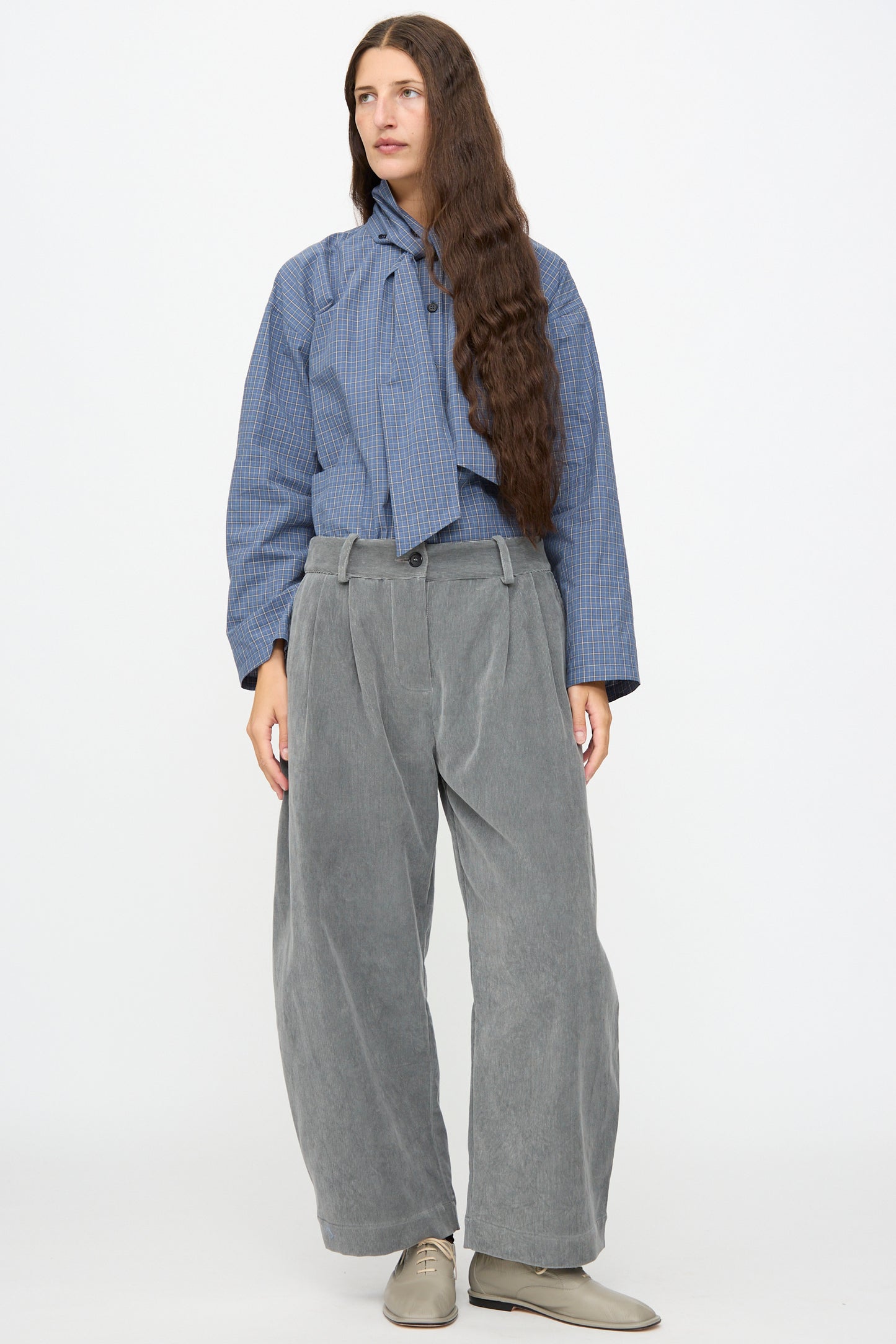 A person stands against a plain background, wearing a blue checkered blouse with a neck tie, Cawley’s Japanese Wale Cotton Cord Robyn Trouser in Fossil featuring a pleated front and wide legs, paired with light gray shoes.