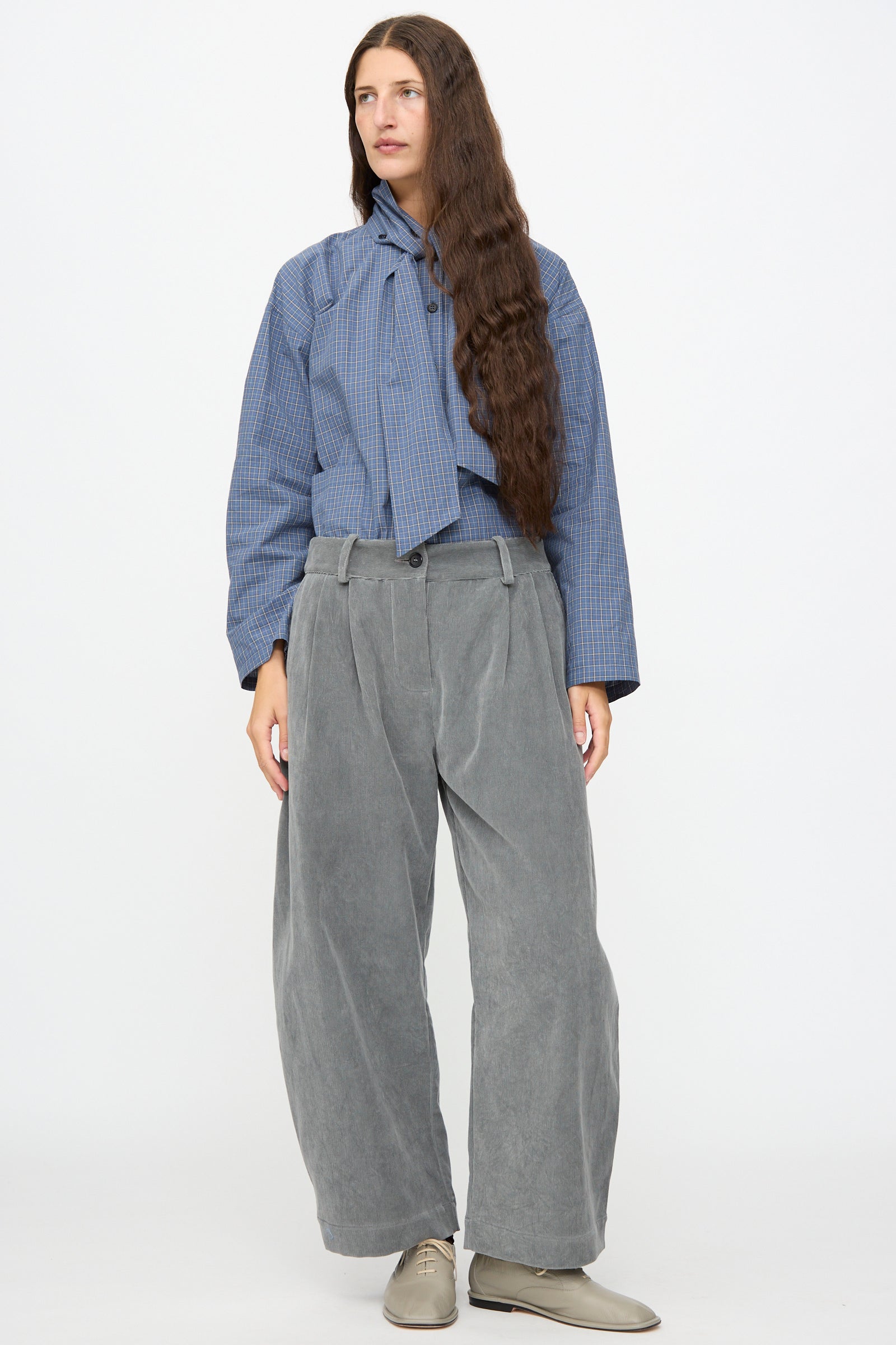 A person stands against a plain background, wearing a blue checkered blouse with a neck tie, Cawley’s Japanese Wale Cotton Cord Robyn Trouser in Fossil featuring a pleated front and wide legs, paired with light gray shoes.