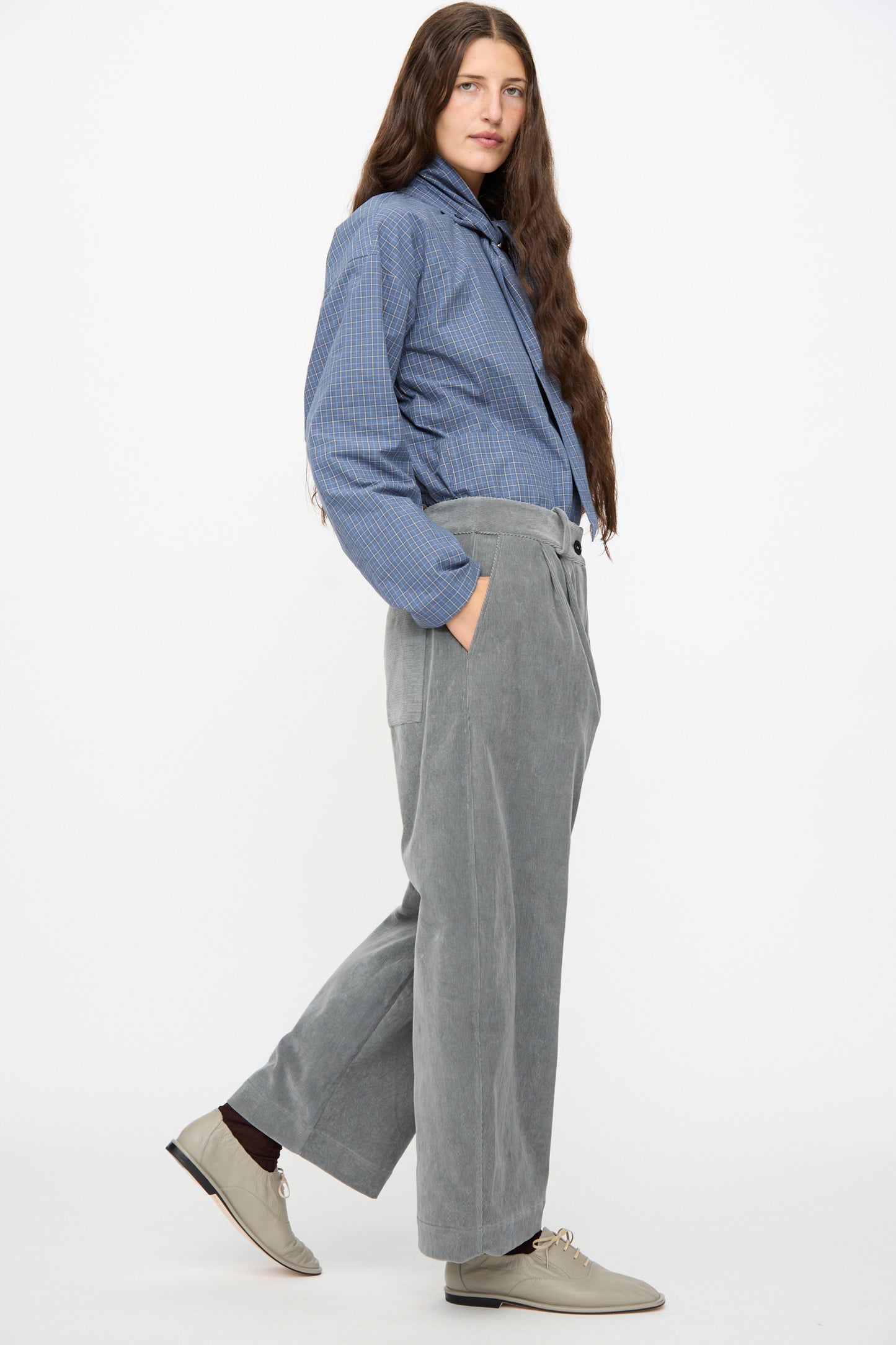 A person with long hair wearing a blue plaid shirt, the Cawley Japanese Wale Cotton Cord Robyn Trouser in Fossil, and beige shoes stands against a plain white background.