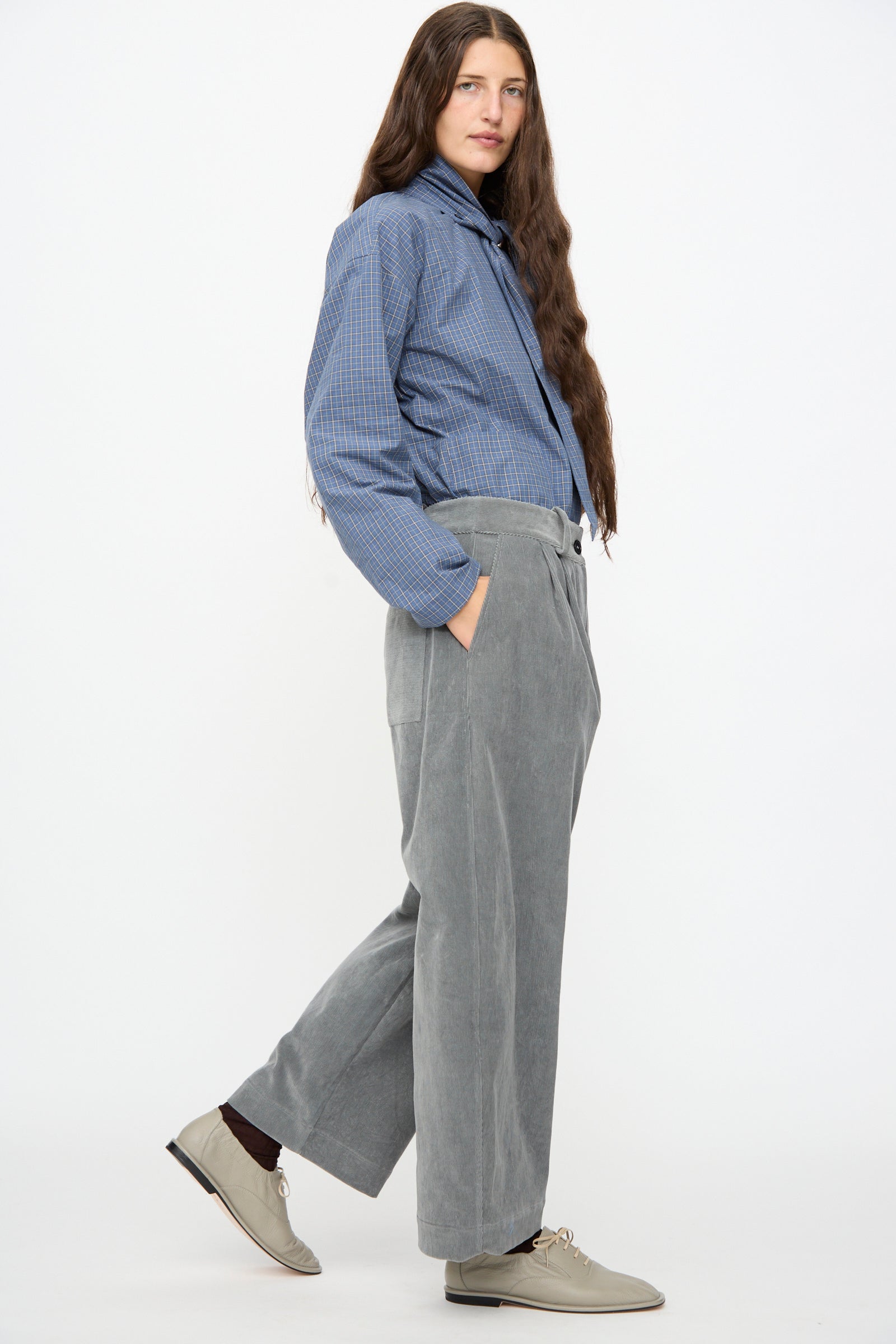 A person with long hair wearing a blue plaid shirt, the Cawley Japanese Wale Cotton Cord Robyn Trouser in Fossil, and beige shoes stands against a plain white background.