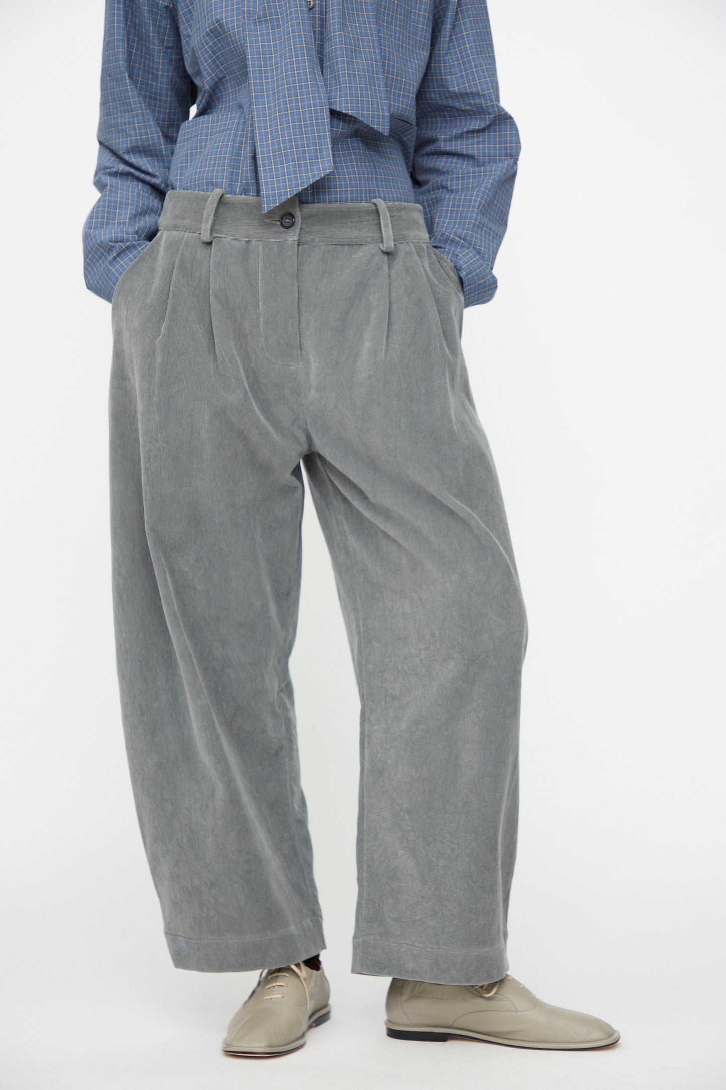 A person is wearing Cawley’s Japanese Wale Cotton Cord Robyn Trouser in Fossil, styled with beige shoes and a blue checkered shirt. Their hands are in their pockets, with only the lower half of the body visible.