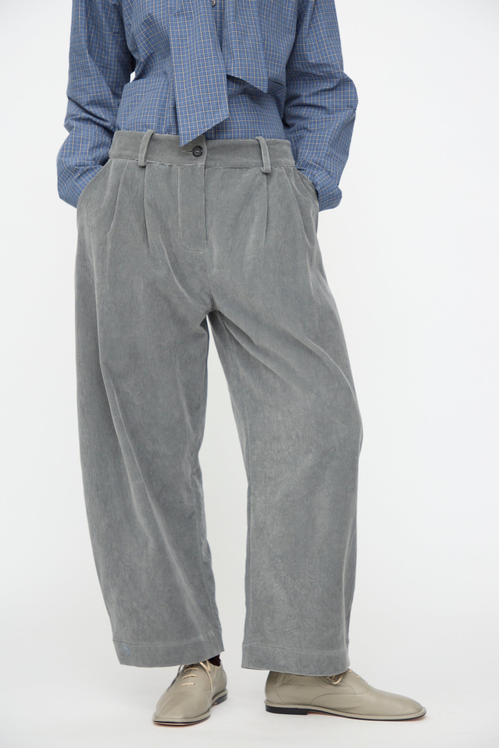 A person is wearing Cawley’s Japanese Wale Cotton Cord Robyn Trouser in Fossil, styled with beige shoes and a blue checkered shirt. Their hands are in their pockets, with only the lower half of the body visible.