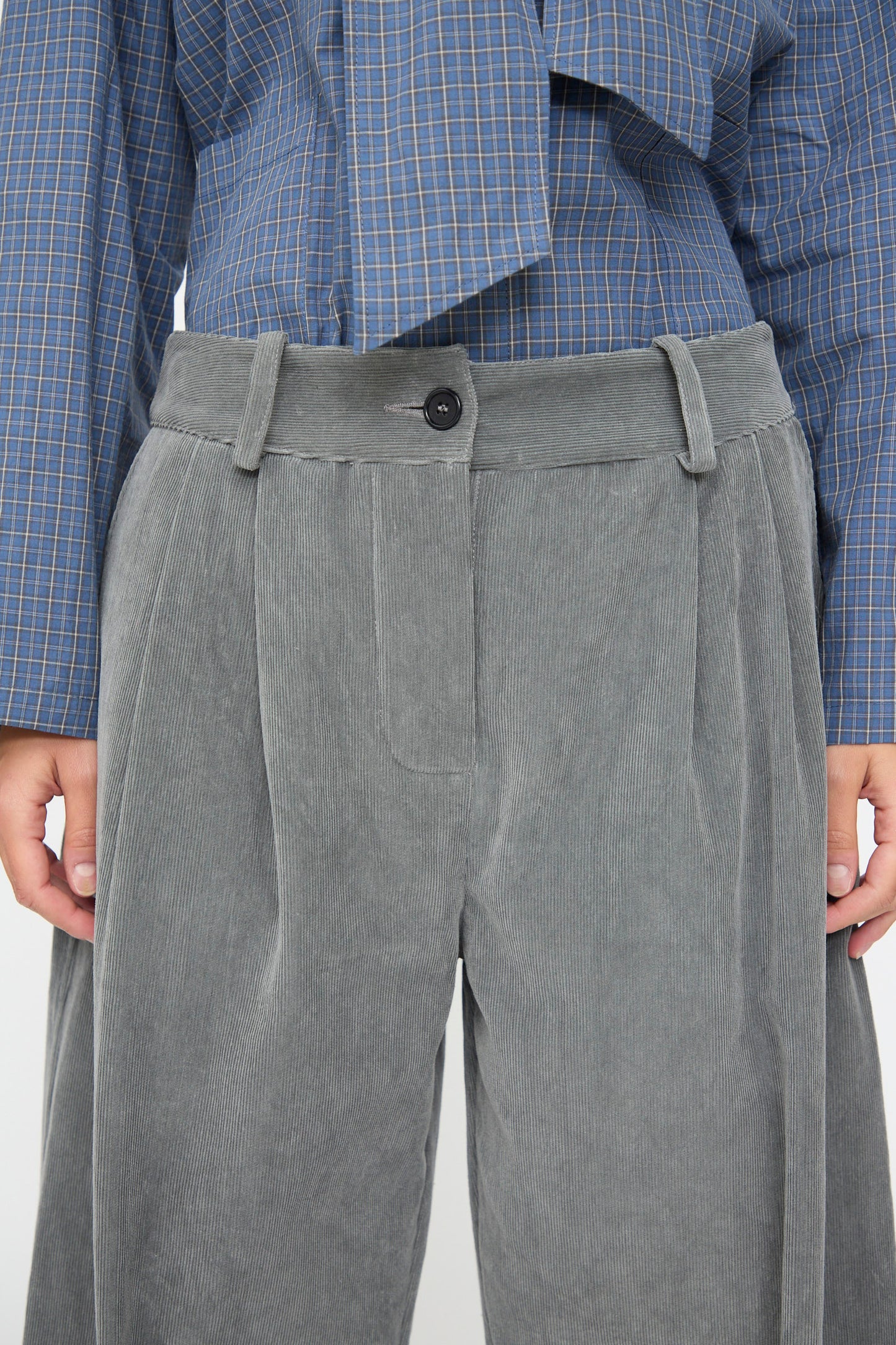 A person is wearing the Cawley Japanese Wale Cotton Cord Robyn Trouser in Fossil, featuring a wide-leg silhouette, paired with a blue plaid shirt; shown from mid-torso to mid-thigh against a plain background.