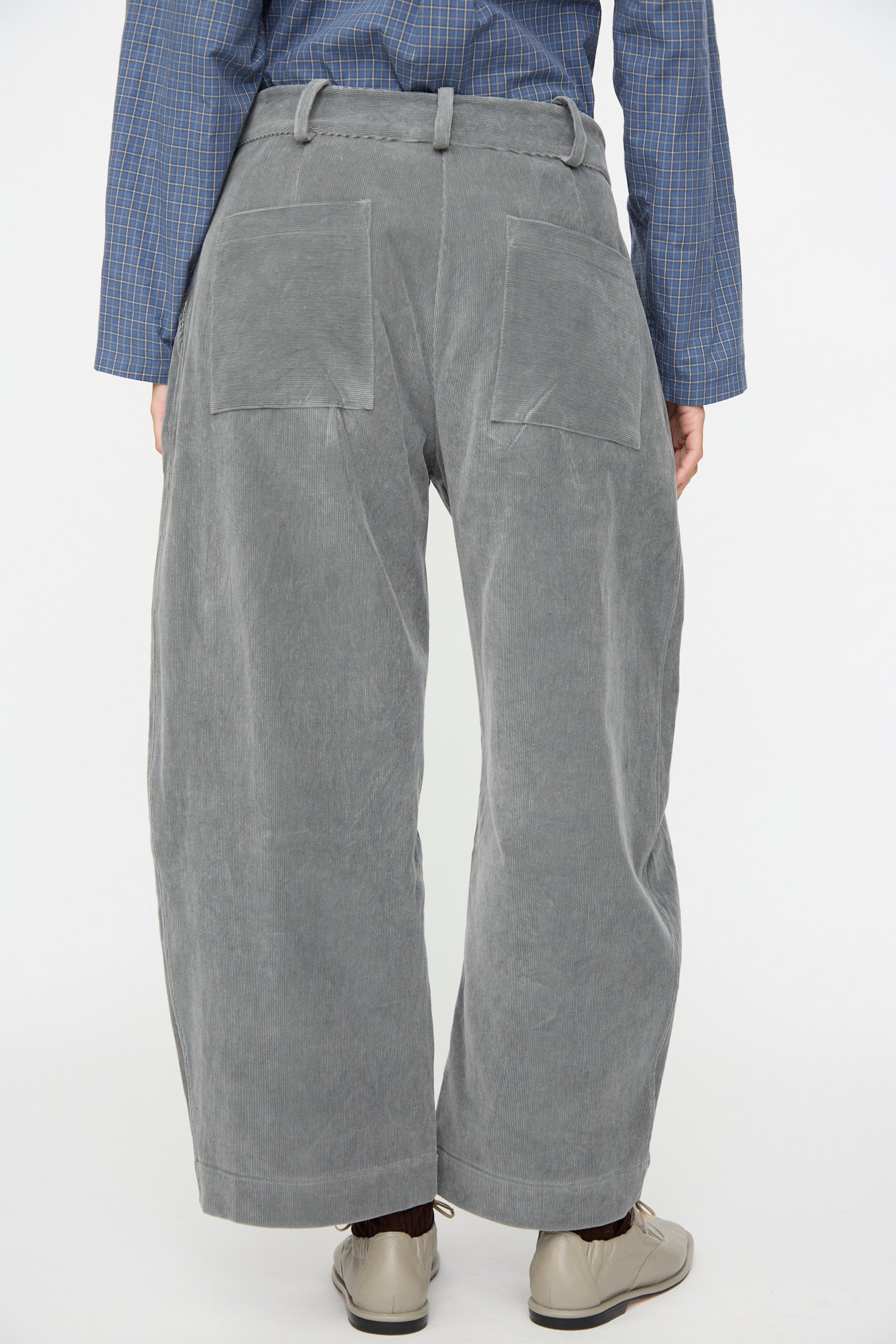 A person is shown from the back against a plain white background, wearing the Cawley Japanese Wale Cotton Cord Robyn Trouser in Fossil, featuring large back pockets, paired with a blue plaid shirt and beige shoes.