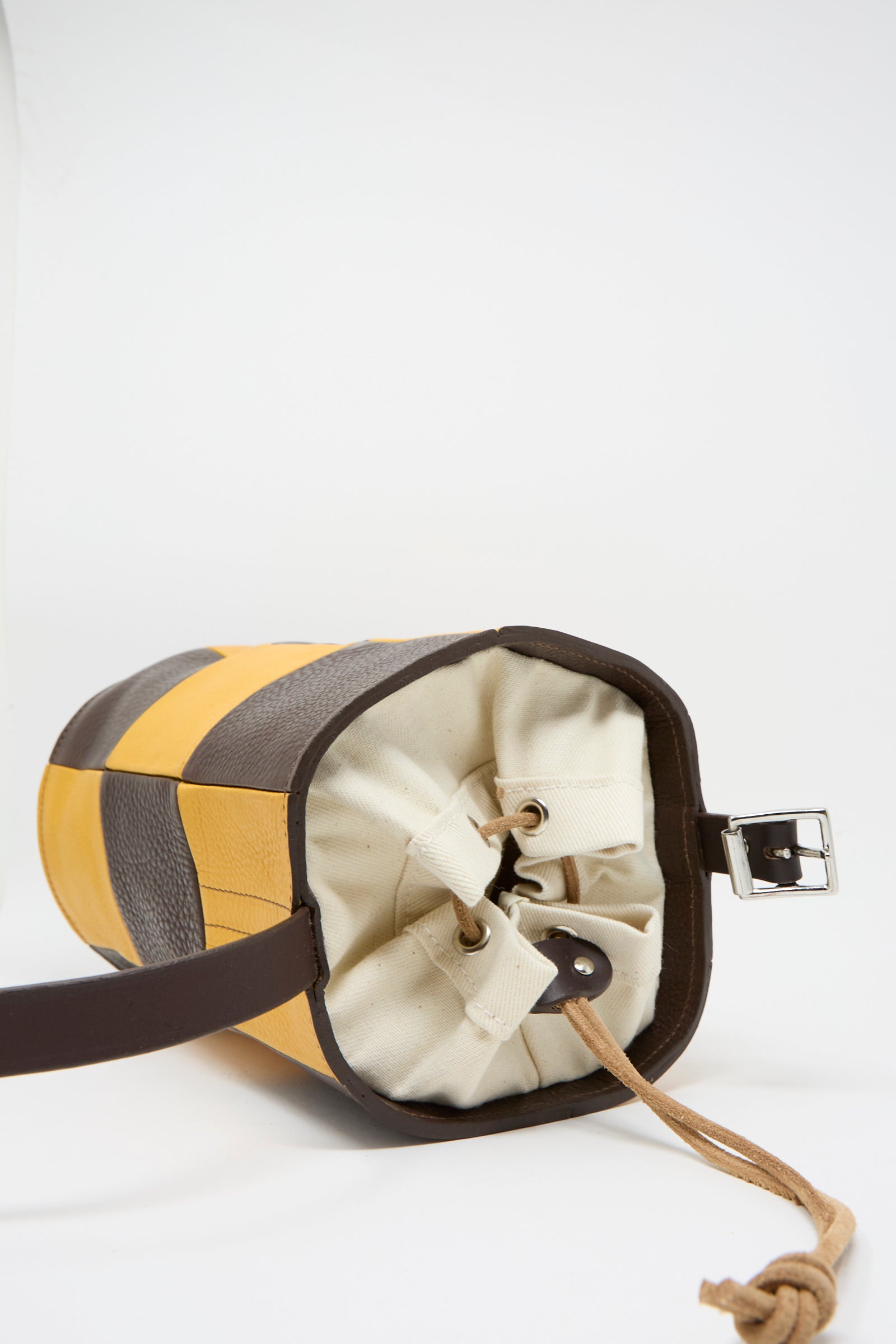 The Cawley Leather Patchwork Bucket Bag in Tan and Mustard is a stylish cylindrical piece with yellow, gray, and cream stripes, elegant brown trim, and a dark adjustable strap, adding versatility to its design.