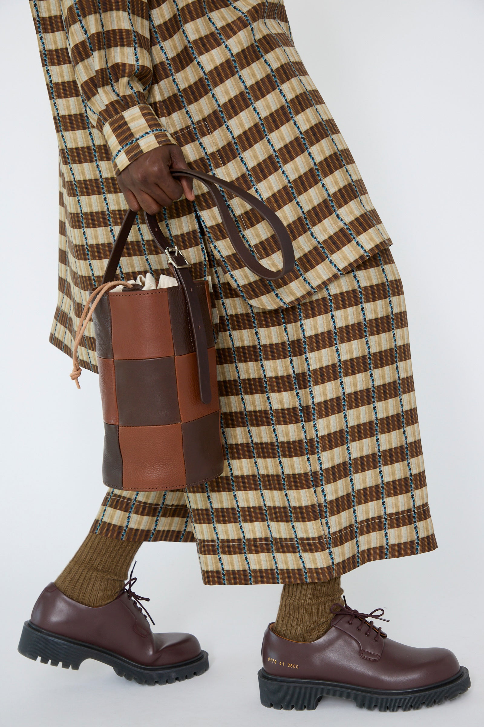 A person dressed in a brown checkered outfit and shoes carries the Leather Bucket Bag in Chocolate and Tan from Cawley, which features an adjustable strap.