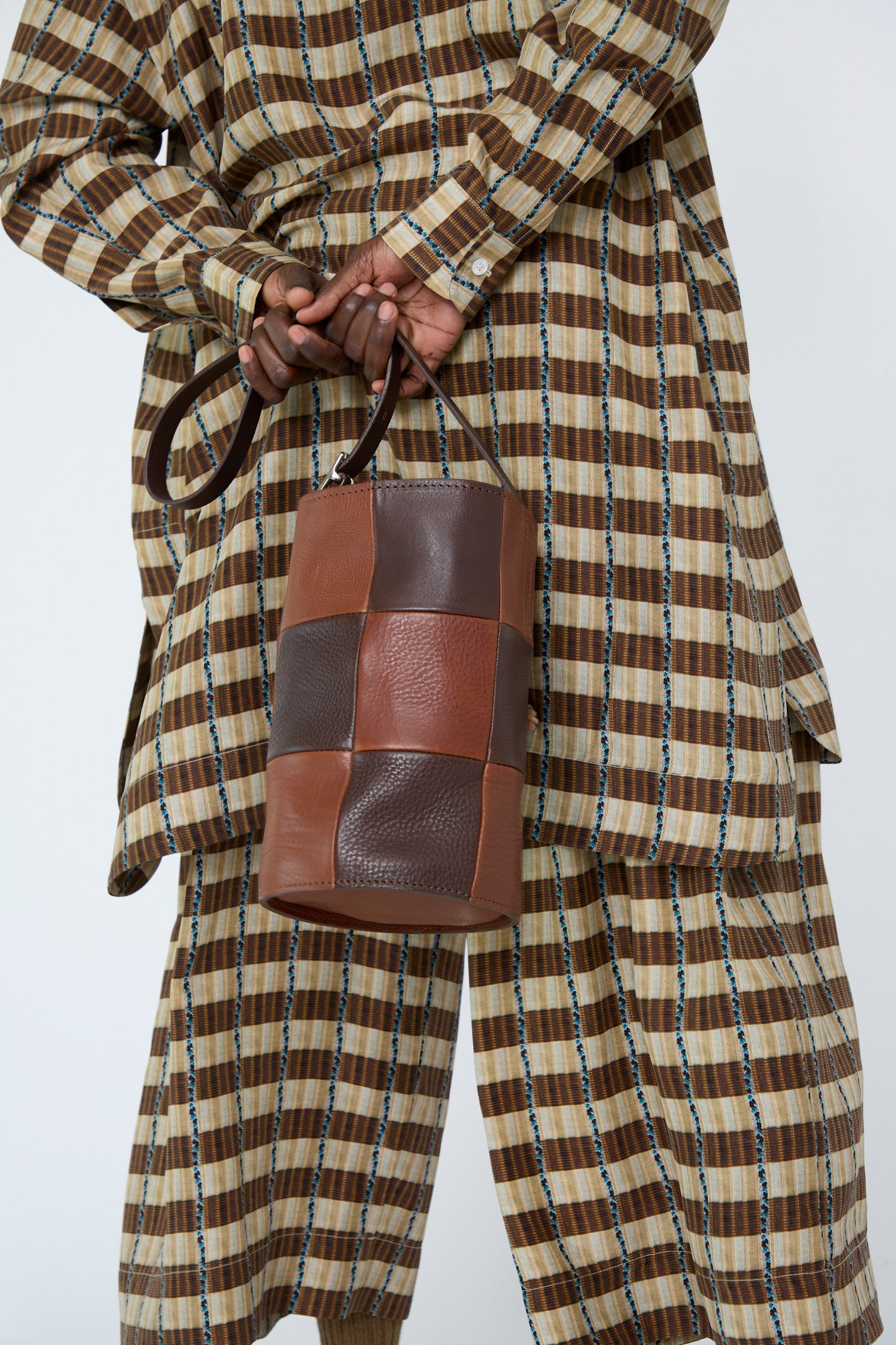 A woman in a checkered outfit holding the Leather Bucket Bag in Chocolate and Tan by Cawley, below the shoulders.