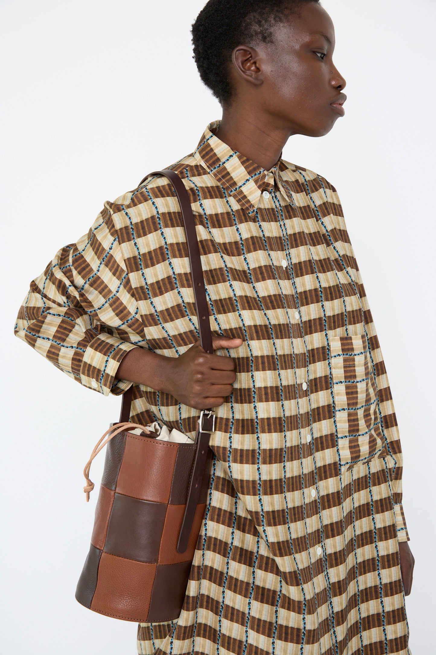 A woman wearing a brown checkered shirt holds a Leather Bucket Bag in Chocolate and Tan by Cawley over her shoulder.