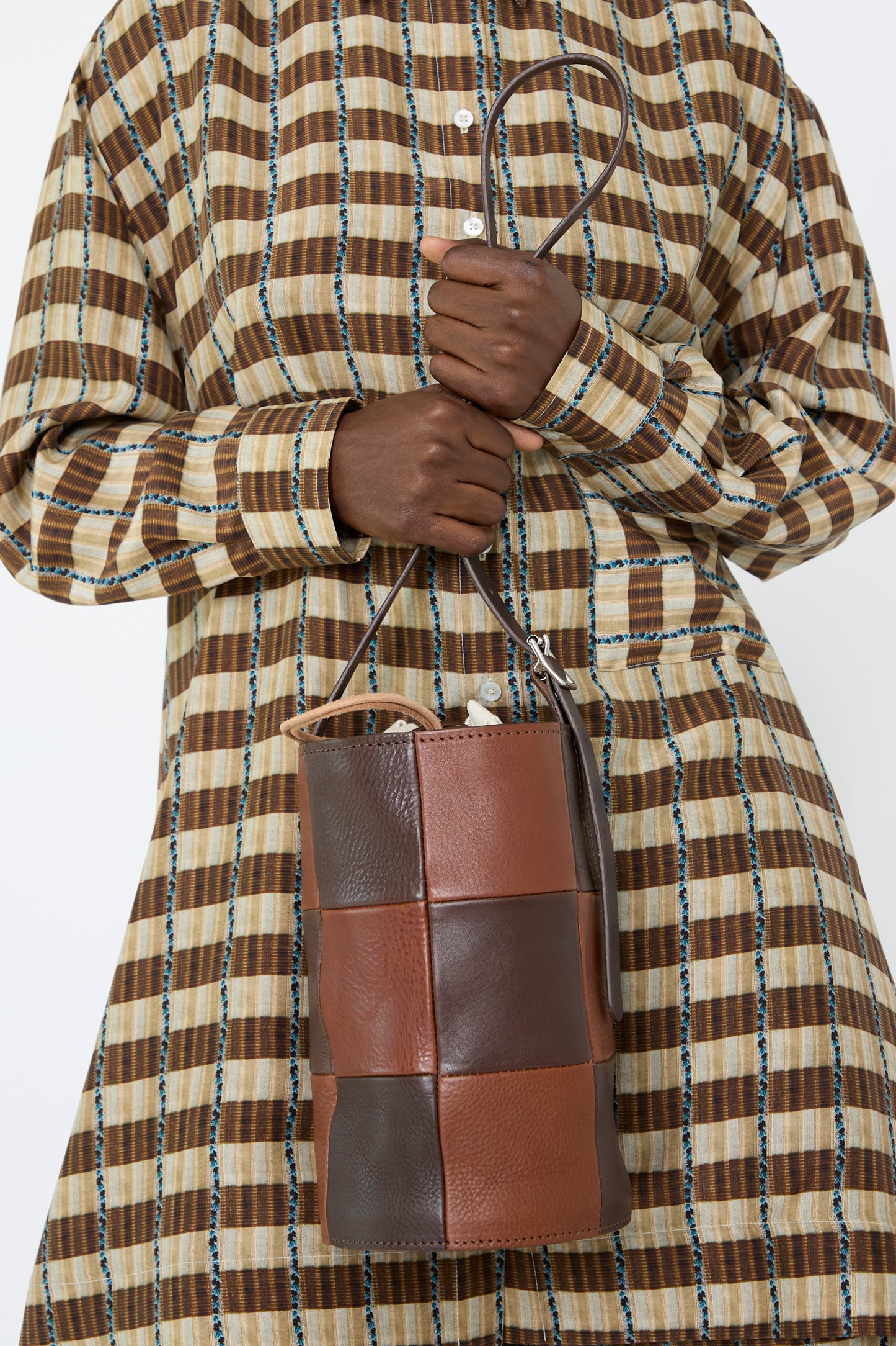 A woman wearing a plaid dress holds a stylish Leather Bucket Bag in Chocolate and Tan made by Cawley, pictured below the shoulder.