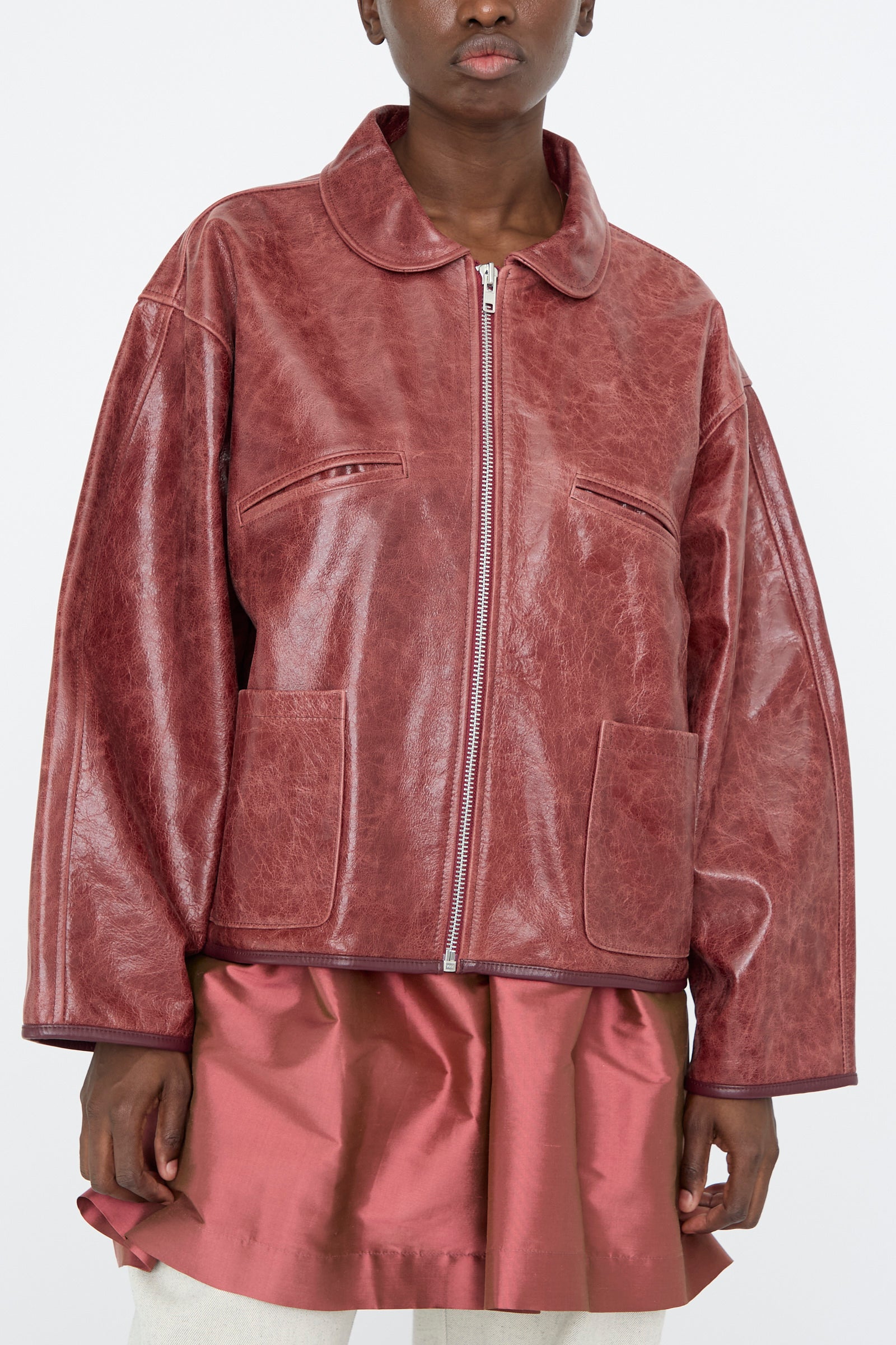 Someone wearing the Cawley Leather Flying Jacket in Burgundy, characterized by its shiny leather, oversized fit, and zip-up front, paired with a matching skirt.