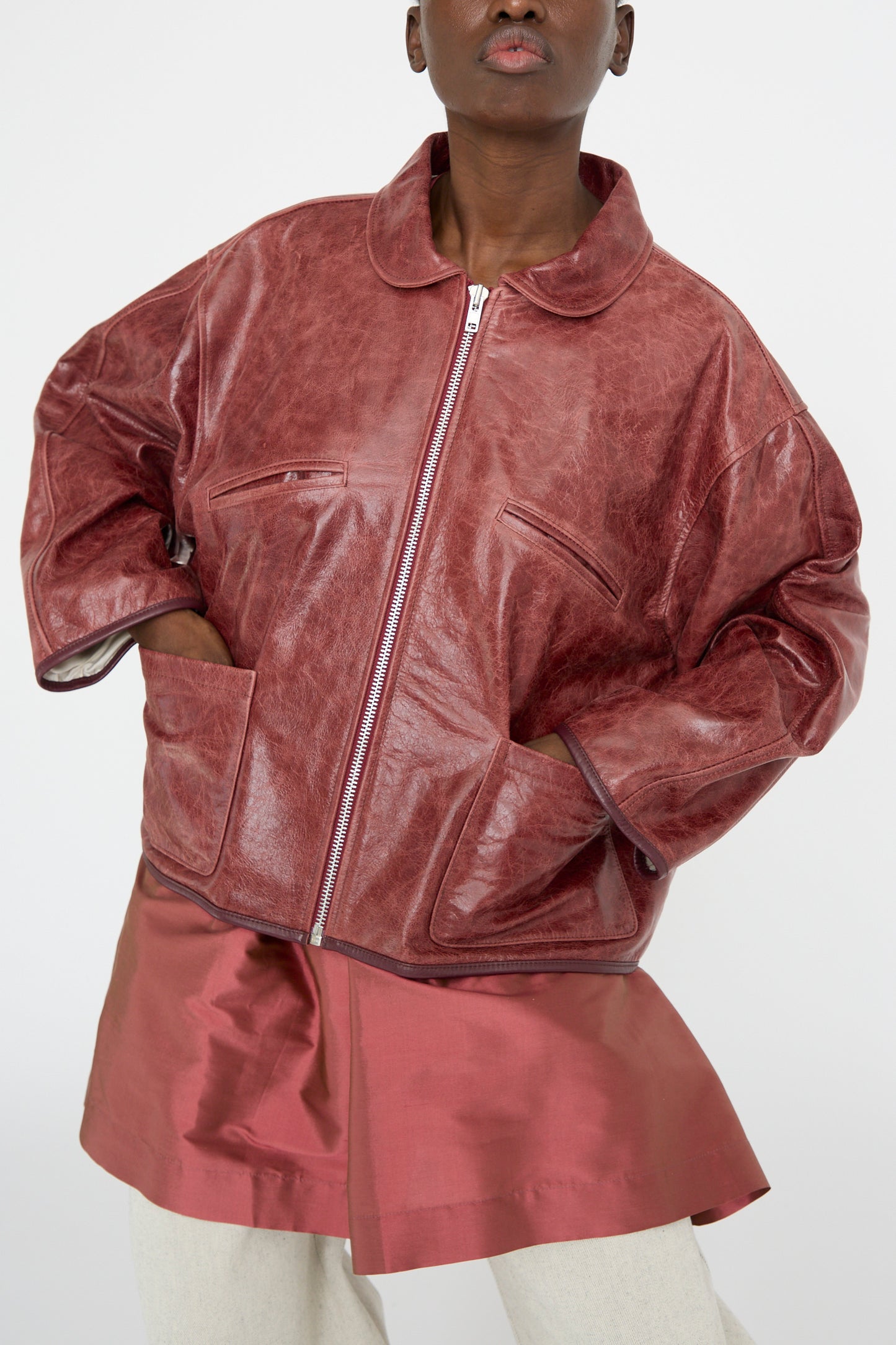 A person models a Cawley Leather Flying Jacket in Burgundy with a matching skirt, hands nonchalantly tucked in pockets, set against a plain white background.