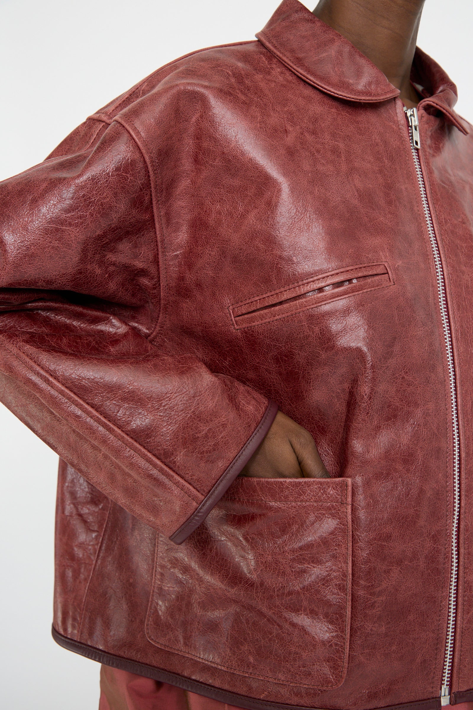 A person wears the Leather Flying Jacket in Burgundy by Cawley, with hands in pockets. The jacket includes a zipper and a small breast pocket.