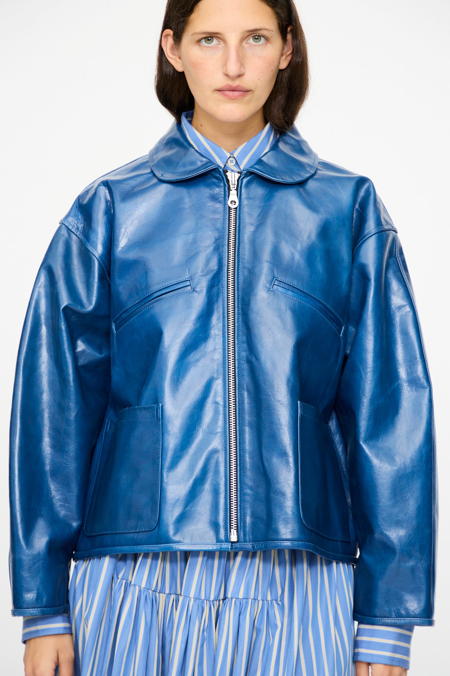 A person wearing the Cawley Leather Flying Jacket in Canada Blue, styled with a blue and white striped collared shirt and pleated skirt, stands against a plain background.