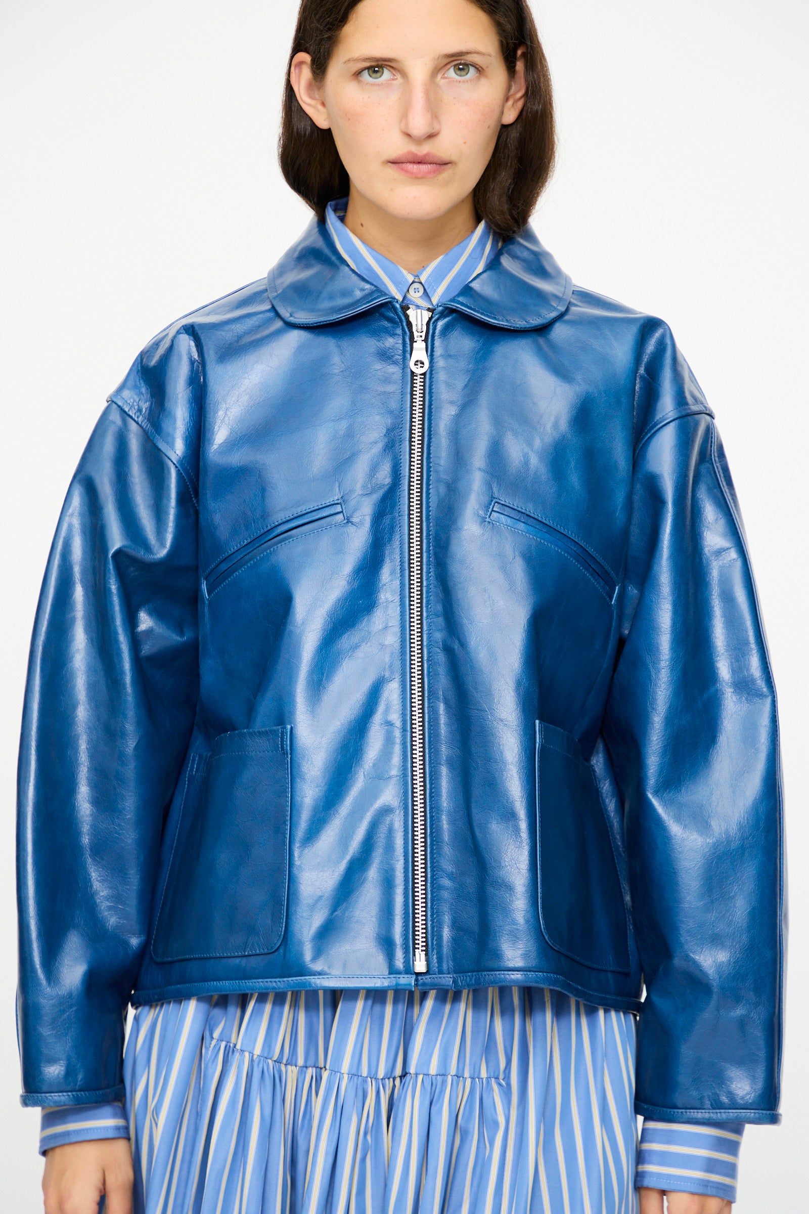 A person wearing the Cawley Leather Flying Jacket in Canada Blue, styled with a blue and white striped collared shirt and pleated skirt, stands against a plain background.