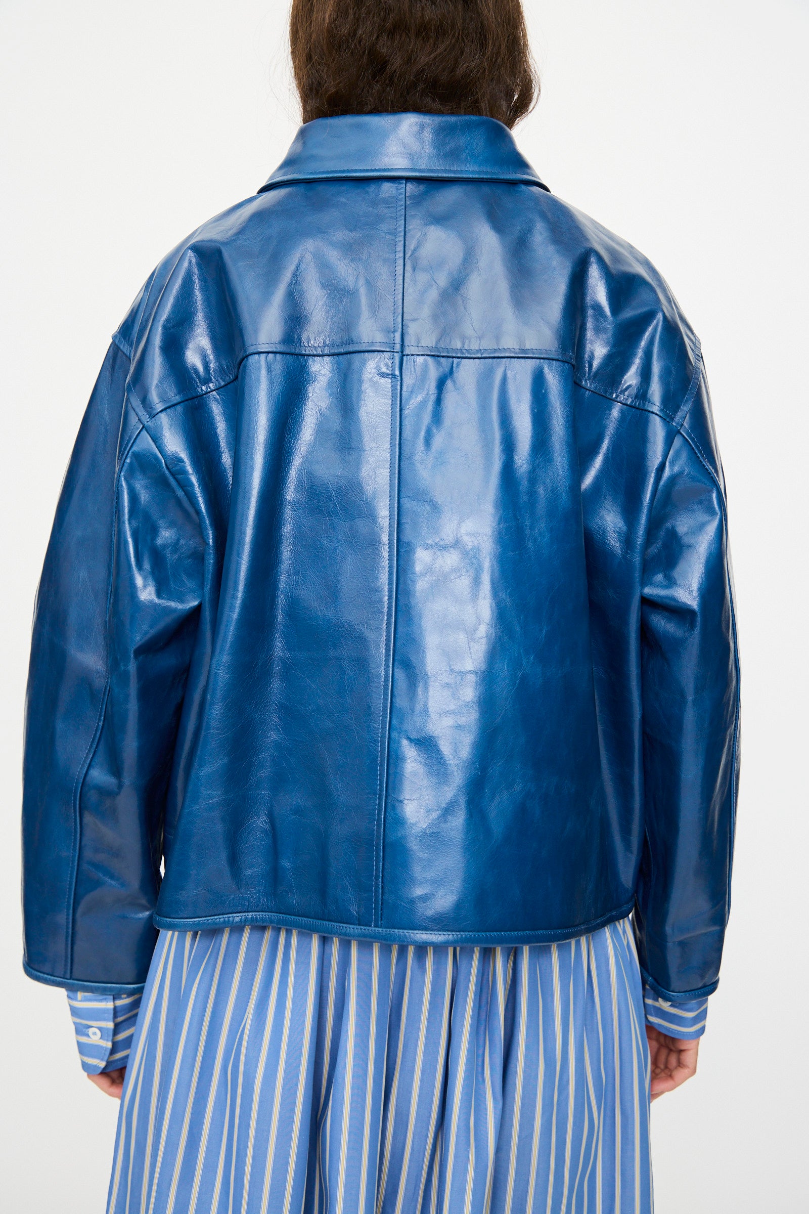 A person wears the Cawley Leather Flying Jacket in Canada Blue, featuring an oversized fit, layered over a blue and white striped dress, photographed from behind against a plain background.