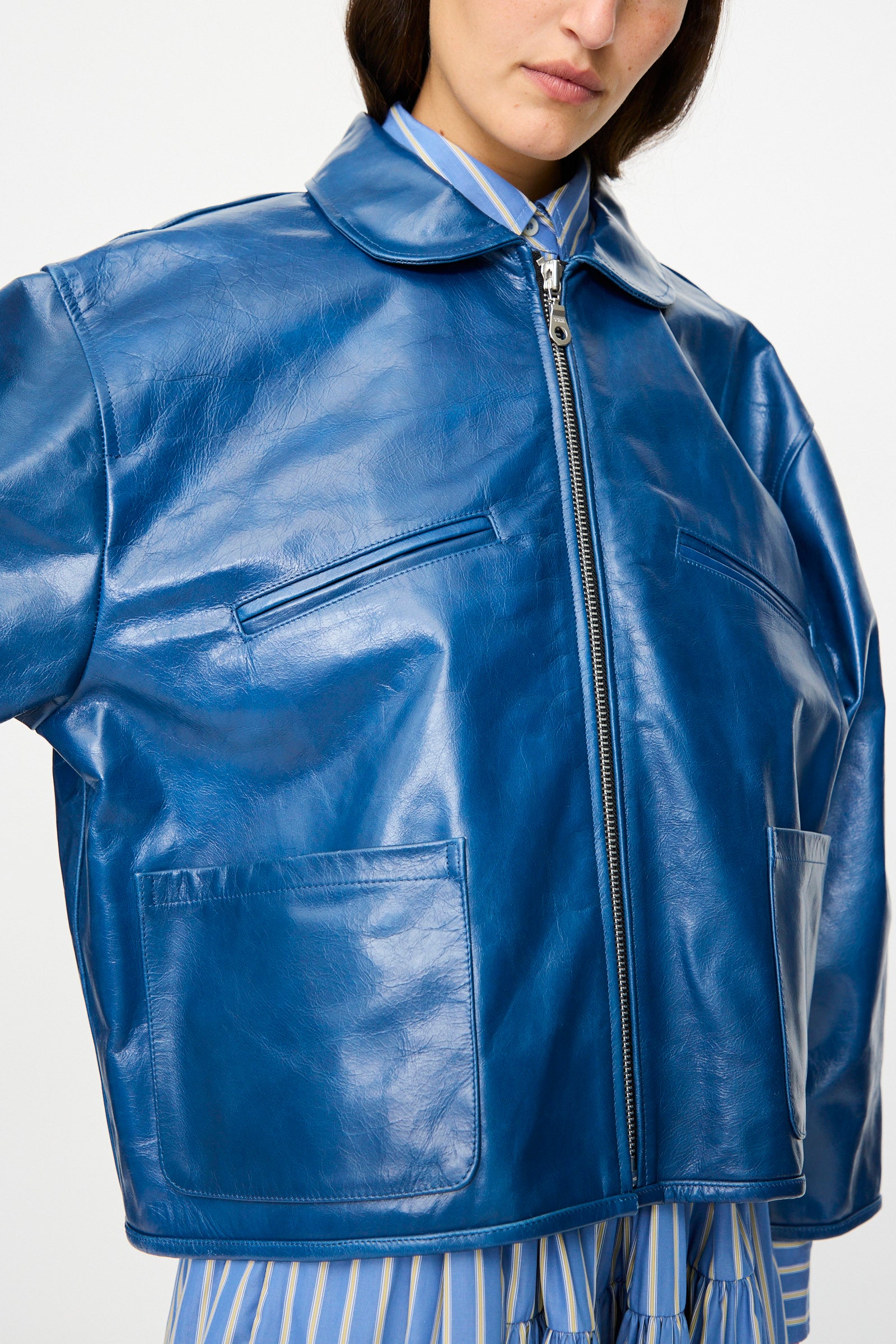 A person wears the Cawley Leather Flying Jacket in Canada Blue—an oversized, shiny blue aviator jacket with front zipper, large collar, chest and lower pockets—styled over a blue and yellow striped shirt.
