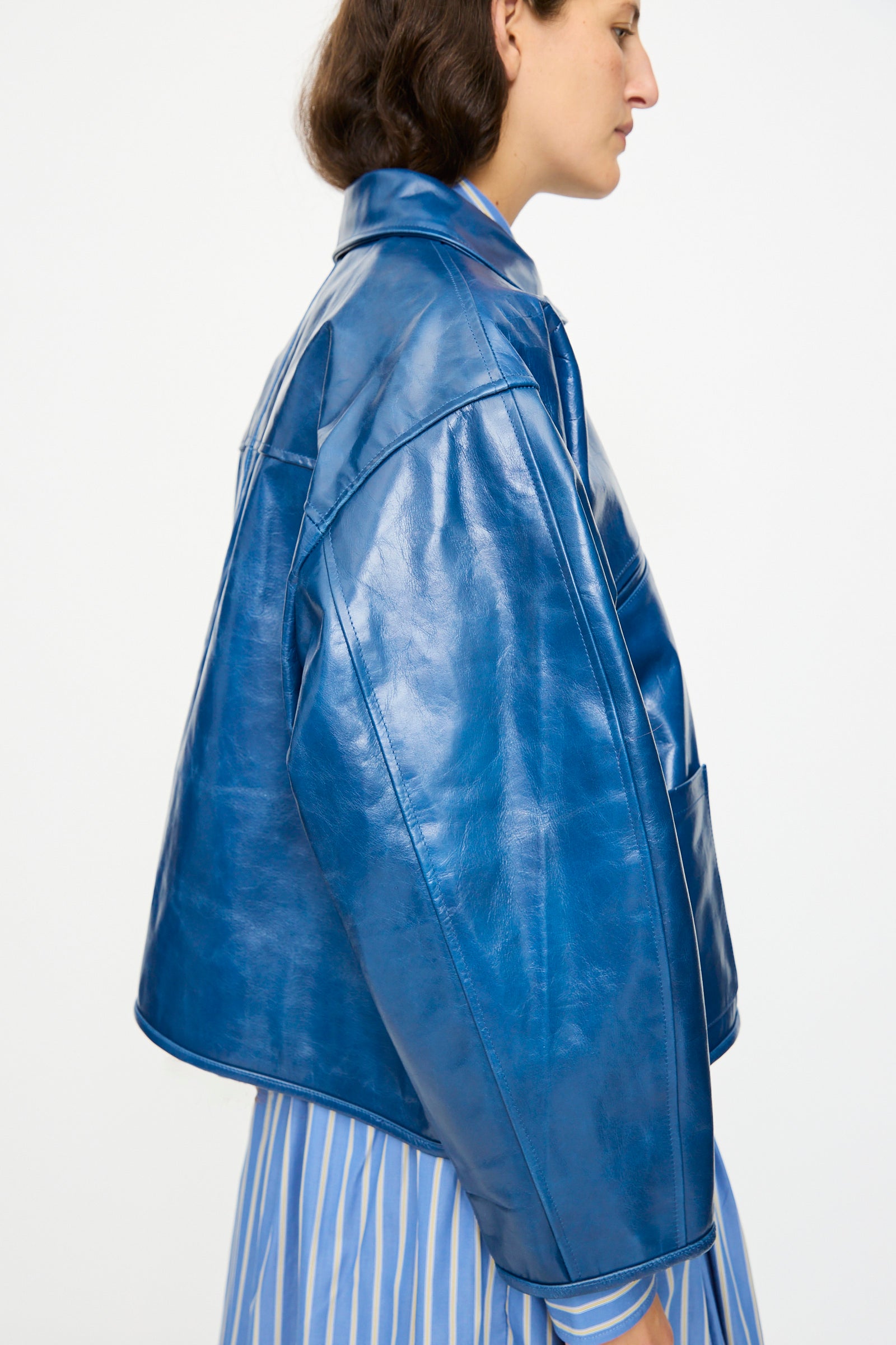 A person in profile wears the Cawley Leather Flying Jacket in Canada Blue—an oversized, shiny blue aviator style—over a blue and white striped shirt, set against a plain light background.