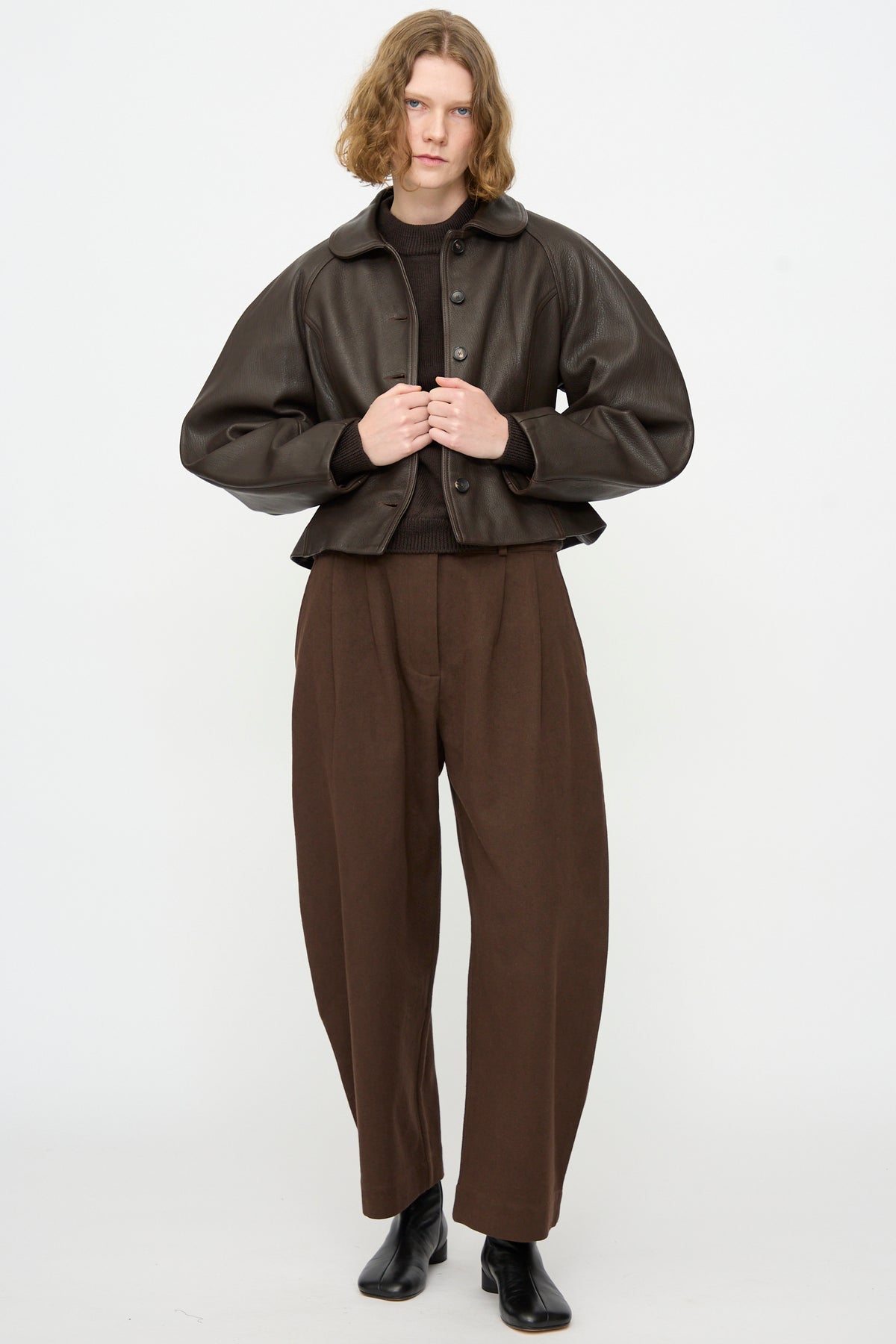 A person stands against a white background, wearing Cawley’s Leather Heidi Boned Jacket in Brown, paired with brown wide-leg trousers, a brown sweater, and black ankle boots.