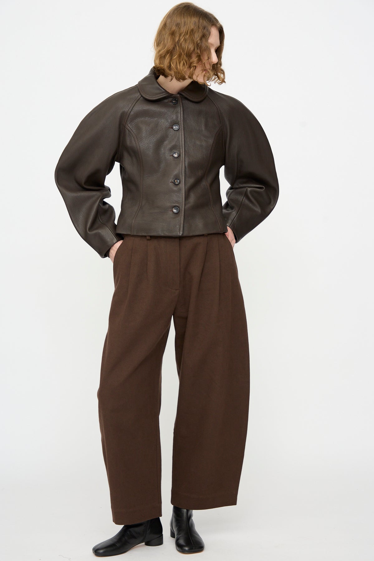 A person stands against a plain white background, hands in pockets and looking to the side, wearing Cawley’s Leather Heidi Boned Jacket in Brown styled with wide brown pants and black boots.