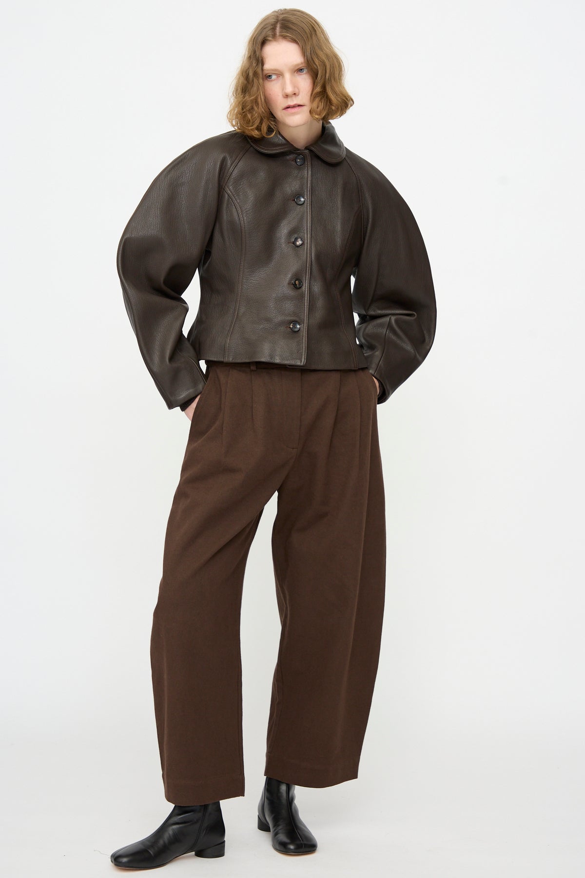 A person stands against a plain background wearing the Cawley Leather Heidi Boned Jacket in Brown, paired with wide-leg brown pants and black ankle boots, hands in pockets and a neutral expression.