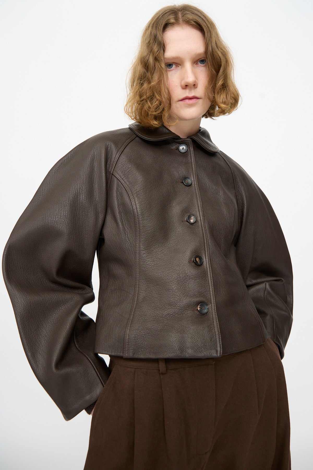 A person wearing the Cawley Leather Heidi Boned Jacket in Brown—a dark brown, sculptural jacket with wide sleeves crafted from goat leather—paired with brown trousers, standing against a plain white background.