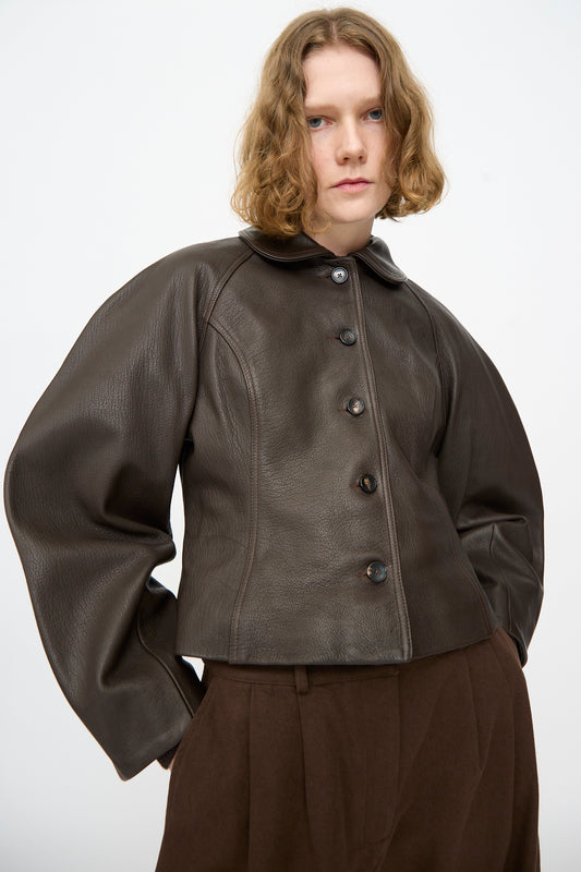 A person wearing the Cawley Leather Heidi Boned Jacket in Brown—a dark brown, sculptural jacket with wide sleeves crafted from goat leather—paired with brown trousers, standing against a plain white background.