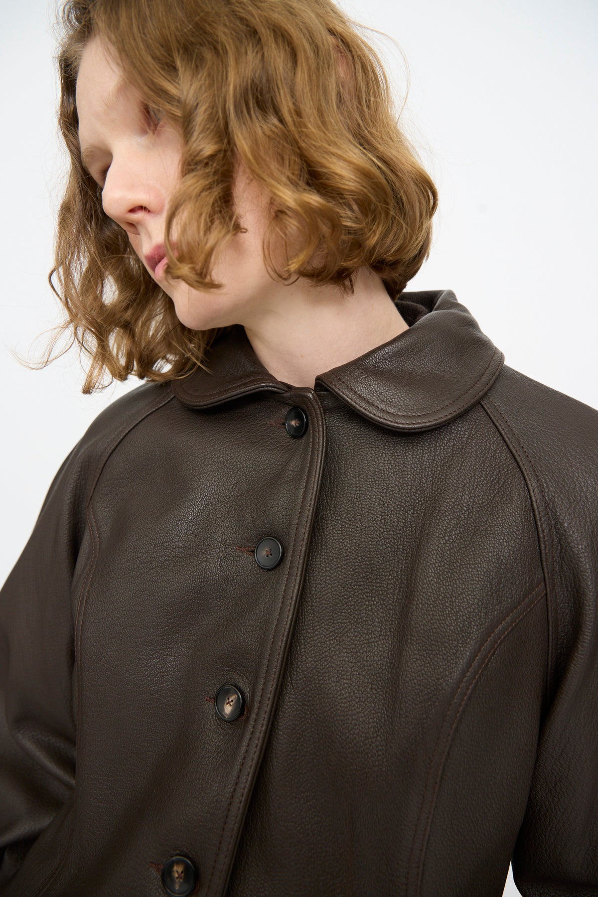 A person with wavy, shoulder-length hair wears the Cawley Leather Heidi Boned Jacket in Brown—a dark brown goat leather button-up coat with a rounded collar—standing against a plain light background.