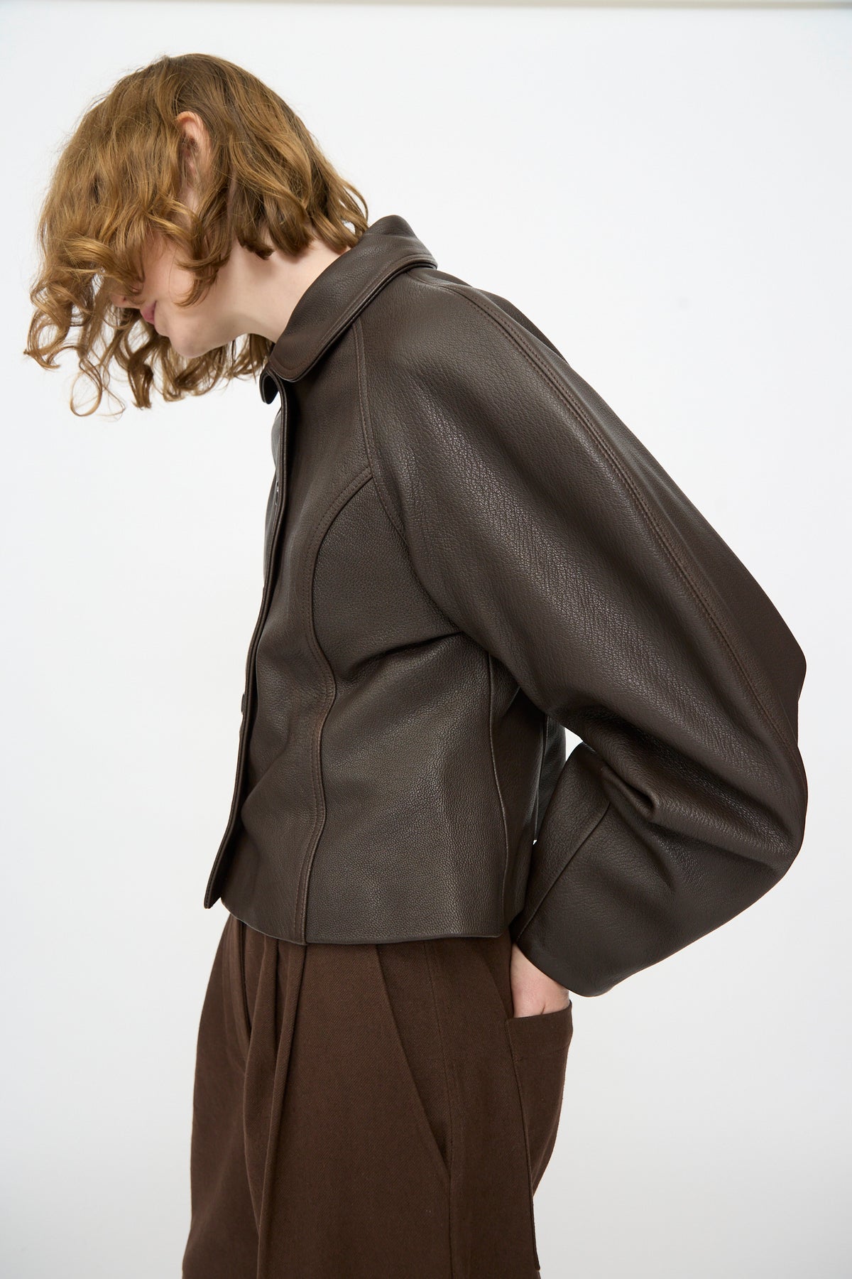 A person with wavy light brown hair wearing the Cawley Leather Heidi Boned Jacket in Brown and brown pants, standing in profile against a plain white background.