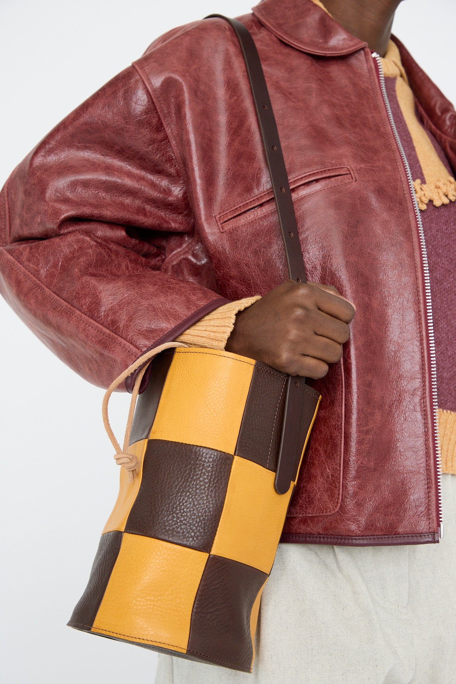 A person in a red leather jacket holds a Cawley Leather Patchwork Bucket Bag in Tan and Mustard, featuring an adjustable strap.