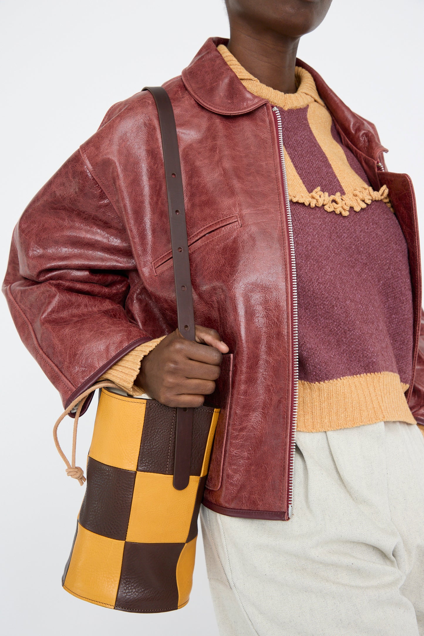 A person in a red leather jacket, maroon and orange sweater, beige pants, holding a Cawley Leather Patchwork Bucket Bag in Tan and Mustard.